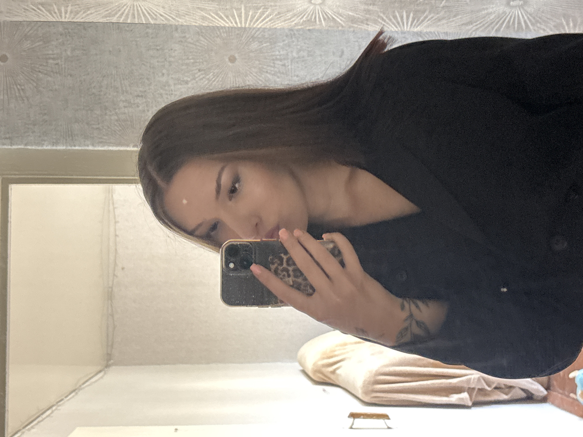 A young woman with long, dark brown hair is taking a mirror selfie in a room. She is wearing a black blazer. Her hair is parted in the middle and falls past her shoulders. Her eyes are open, and she is pursing her lips. She is holding a smartphone with a leopard print case in her right hand, obscuring part of her face. She has a tattoo on her left forearm. In the background, a beige garment is draped over an object on the left side of the image, and textured wallpaper with a sunburst pattern is visible on the right. The environment appears to be an indoor residential setting, possibly a bedroom or dressing area. There is no discernible indication of the city of Inskoy, Russia.