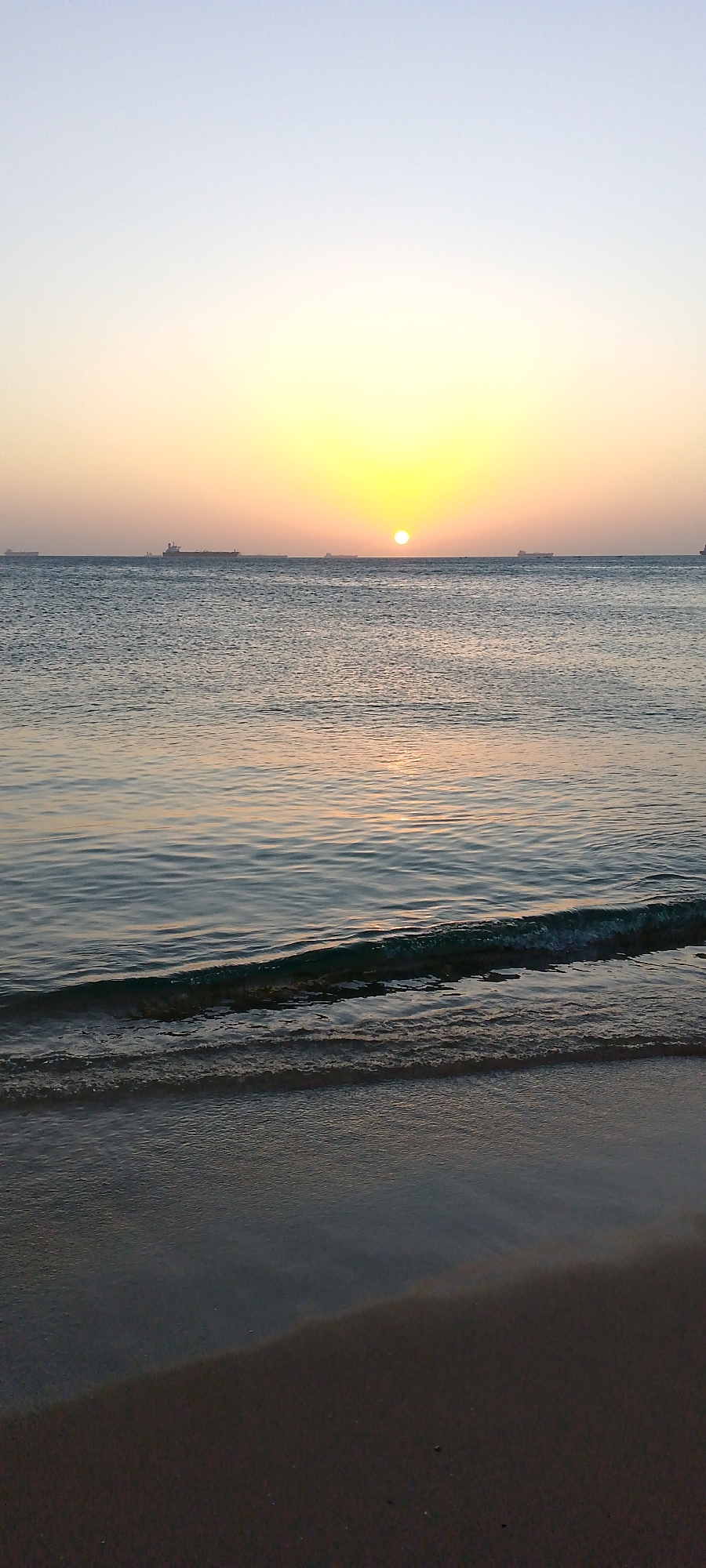 A calm beach at sunset in Villa Marina, Venezuela. The sun is setting over the horizon, with several ships visible in the distance. Gentle waves are lapping at the sandy shore. The sky transitions from light blue at the top to a yellow-orange near the horizon.