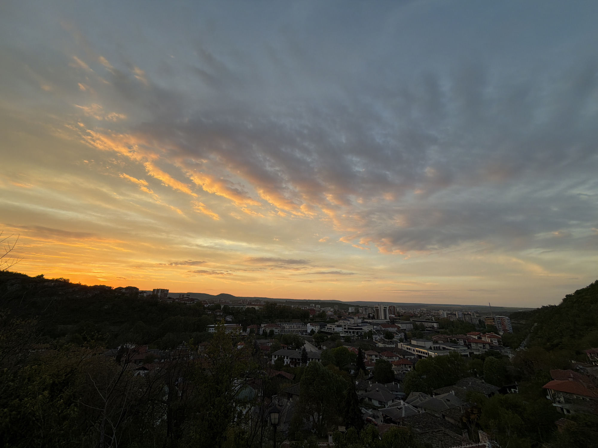 The image captures a scenic view of Lovech, Bulgaria, at dusk or dawn. The sky is a mix of colors, transitioning from a bright orange near the horizon to a softer pink and gray higher up, with wispy cloud formations spread across the upper portion of the frame.
Below the horizon, a city landscape unfolds. There's a dense area of trees and greenery on the left, suggesting a park or forest edge. Residential buildings with visible roofs are interspersed among the trees, leading to more densely packed buildings further into the cityscape. Several taller structures can be seen in the mid-ground, indicating an urban center.
On the far right, another dense patch of trees frames the view, suggesting the photograph was taken from an elevated position overlooking the city. The composition balances the urban and natural elements, with the colorful sky acting as a backdrop.