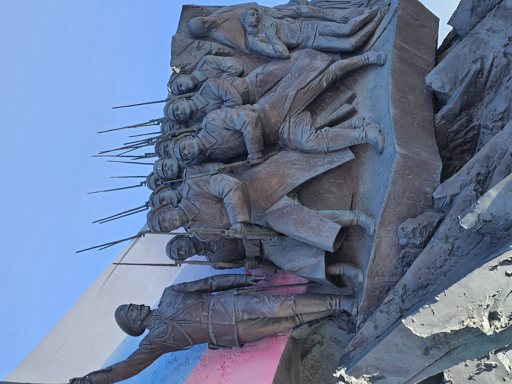 The image shows a section of the Monument to the Heroes of World War I in Moscow, Russia. The bronze relief depicts a group of Russian soldiers advancing with rifles. The sculptures are attached to a stone structure, and the Russian flag is painted below the main figures. The sky is visible in the background.