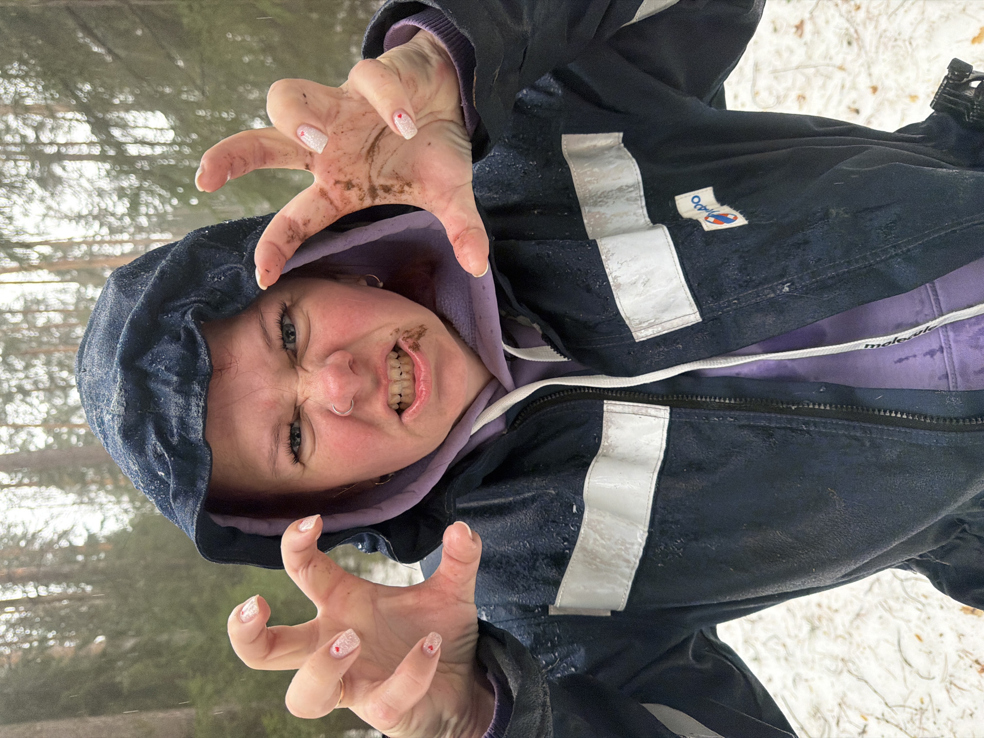 A woman in a navy blue hooded jacket with reflective stripes and a lavender hoodie underneath is posing outdoors in a snowy environment. Her hands are raised with fingers curled like claws, and there's mud or dirt on her hands and around her mouth. She is scowling and her teeth are visible. The background shows trees and snow. The jacket has a small logo on the chest. The specific location in the city of Chaikovsky, Russia, is not identifiable from the image alone.