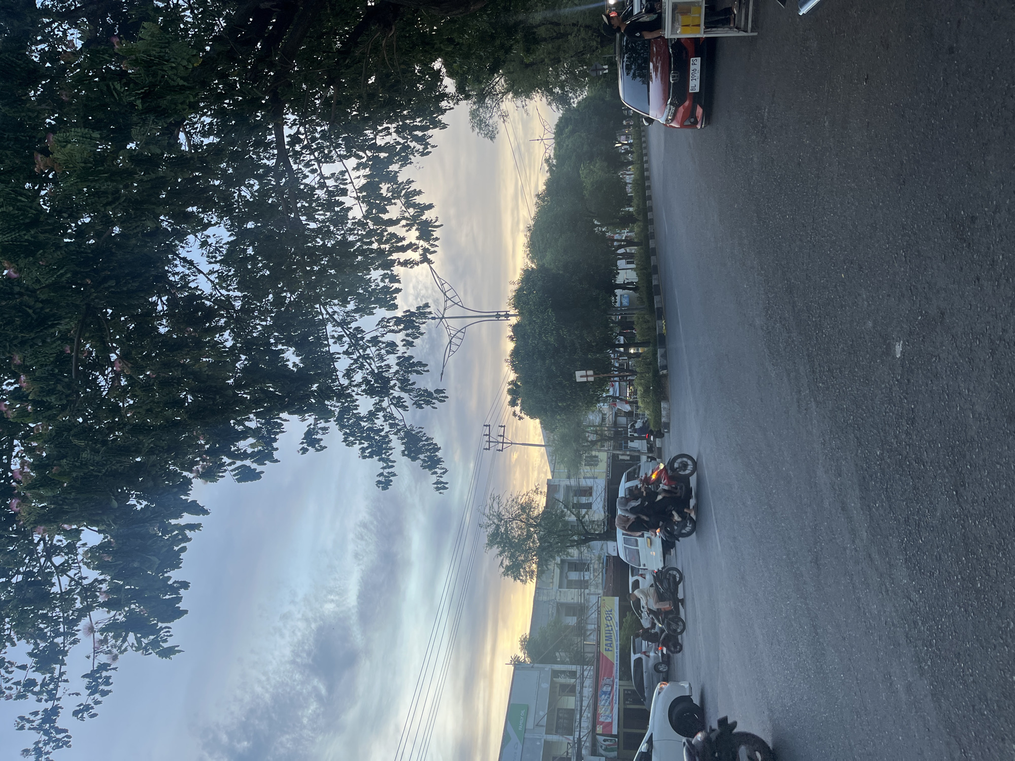 The image depicts a street scene in Banda Aceh, Indonesia, during what appears to be early morning or late afternoon, judging by the soft, diffused light and the sky conditions. The foreground is dominated by a paved road with a slight sheen, indicating recent rain or high humidity. A red car with license plate BL 1996 PS is parked on the right side of the road. To the right of the car is a small, portable food stall with yellow liquids in containers.
In the middle ground, a busy intersection is visible. Several motorcycles are in motion. One motorcycle carries three individuals, two women wearing headscarves and one in the back also appears to be a woman. Another motorcycle in front of it has a man riding it, dressed in light-colored clothing. Further back, other vehicles, including a white van and cars, are present, suggesting traffic flow. Buildings line the left side of the road, some appearing to be shops with signage, including one that reads "FAMILY OIL."
The background features a line of trees forming a natural border along the road. The sky above is a mix of light blue and wispy white clouds, with areas of soft orange and yellow light indicating the position of the sun. Overhead, power lines crisscross the sky, typical of an urban environment. Lush green foliage from a large tree with delicate pink blossoms hangs over the top of the frame, partially obscuring the sky and adding a natural element to the urban landscape. The overall impression is of a dynamic street environment in Banda Aceh, capturing the daily life and infrastructure of the city.