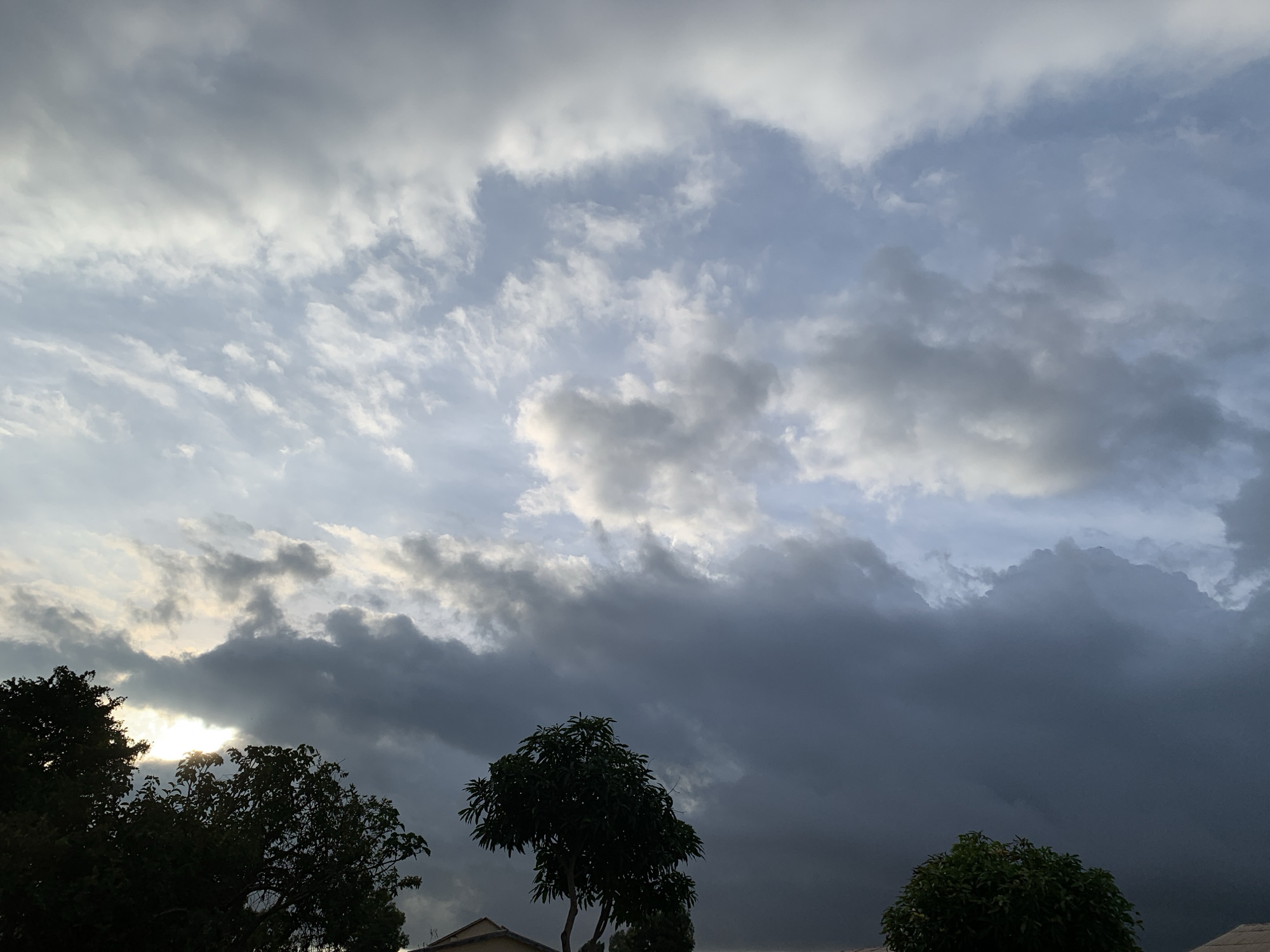 The image depicts a dramatic sky filled with heavy, dark clouds above a silhouette of trees and rooftops in Kuru Karama, Nigeria. The sun is partially visible, casting rays through a break in the clouds on the left side of the frame. The clouds vary in density and color, ranging from lighter grays and whites to deep, ominous blues and blacks, suggesting an approaching storm or the aftermath of one. Silhouetted trees, some appearing to be mango trees, are prominent in the foreground, with their dark foliage contrasting against the turbulent sky. The tops of buildings are visible just above the treeline, hinting at a residential or semi-urban environment. The overall atmosphere is one of natural power and impending change.
