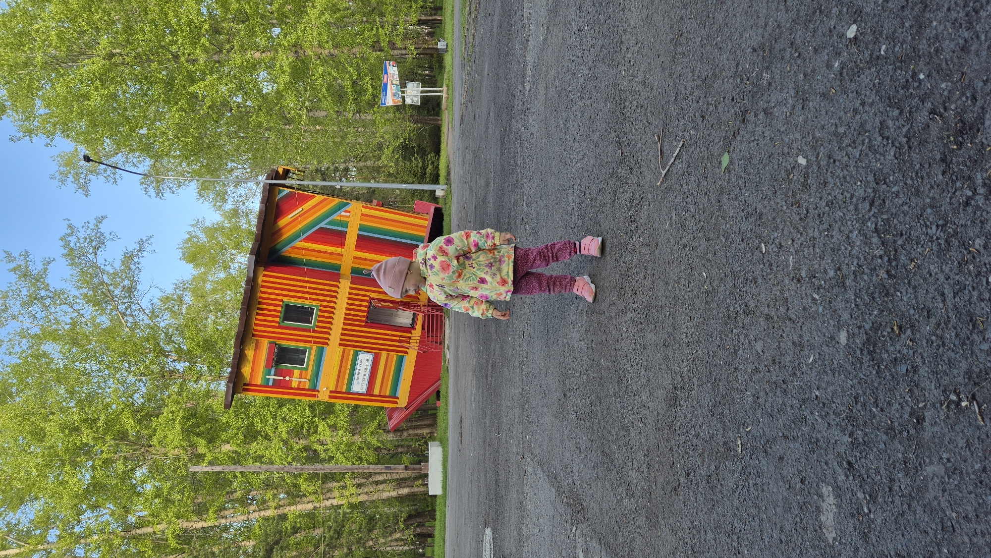 A young child in a pink hat, floral jacket, and pink pants and shoes stands near a brightly colored, two-story building. The building has horizontal stripes of red, orange, yellow, green, blue, and purple. It features multiple windows and a door. The scene is located in Kemerovo, Russia, as the sign on the building displays text in Russian. The child is standing on a paved surface, possibly a road or parking lot. Trees are visible in the background.
