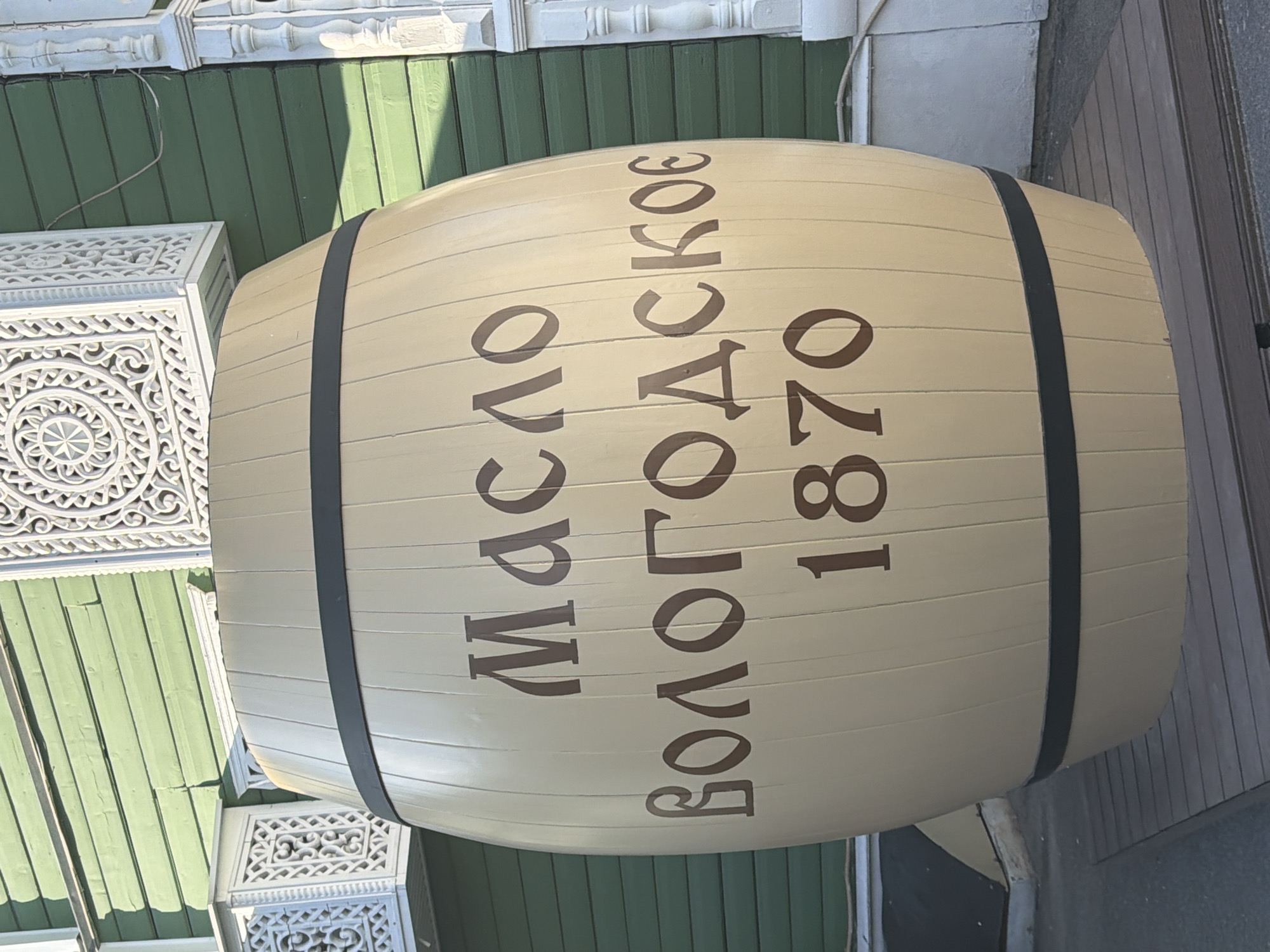 A large, light-colored wooden barrel sits on a wooden platform in front of a green wooden building with white decorative elements. The barrel is emblazoned with dark brown lettering that reads "Масло Вологодское 1870" (Butter Vologodskoye 1870). The building has a green clapboard exterior and white ornate carvings on its balconies and decorative trim. The overall scene appears to be an outdoor display in Vologda, Russia, likely related to the region's famous butter production.
