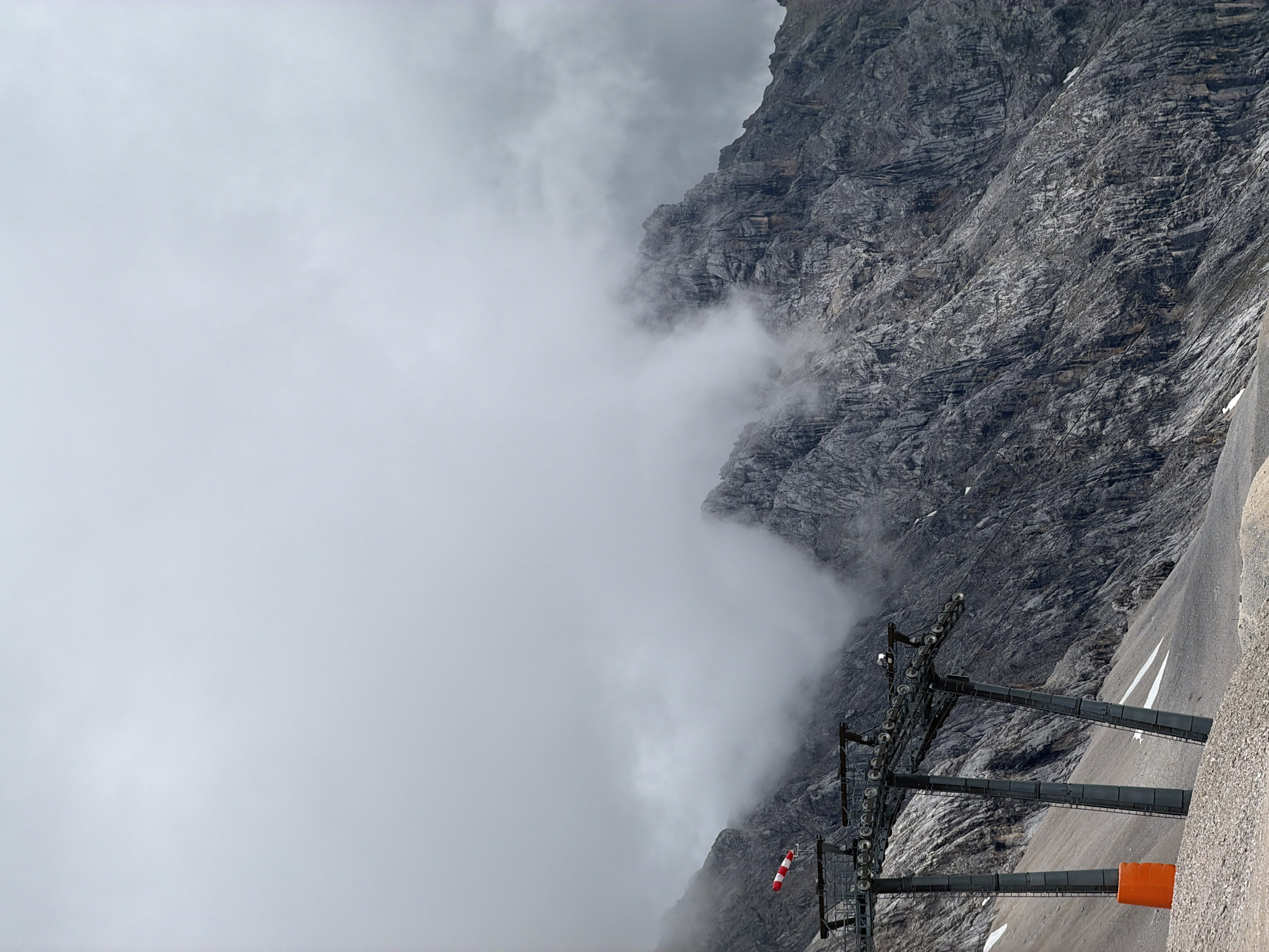 This is a vertical, eye-level, medium shot of a mountain landscape in Garmisch-Partenkirchen, Germany. In the foreground, a ski lift station with three grey support towers and a bright orange cone is visible on a gravelly slope. A red and white windsock hangs from the left tower. The middle and background are dominated by a grey, rocky mountain range partially obscured by thick, white clouds. The sky is overcast and grey. The scene is barren and mountainous, with no people visible.