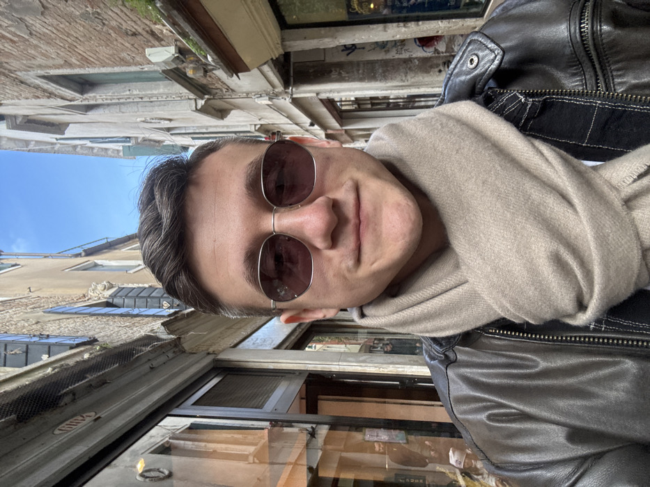 A male individual is depicted in a medium close-up, centered in a narrow Venetian *calle* in Venice, Italy. The subject, appearing to be in his 20s or early 30s, has short dark hair and wears dark oval sunglasses with thin silver frames. He is dressed in a dark brown leather jacket, partially visible, and a light beige woolen scarf wrapped around his neck. His expression is neutral with a faint upward turn at the corners of his mouth. The background features tall, aged stone and stucco buildings characteristic of Venetian architecture, forming the narrow passageway, with a clear blue sky visible directly overhead. On the left, a shopfront with reflective glass and a dark-framed entrance is visible. On the right, another building wall displays some indistinct graffiti and a white wall-mounted fixture.
