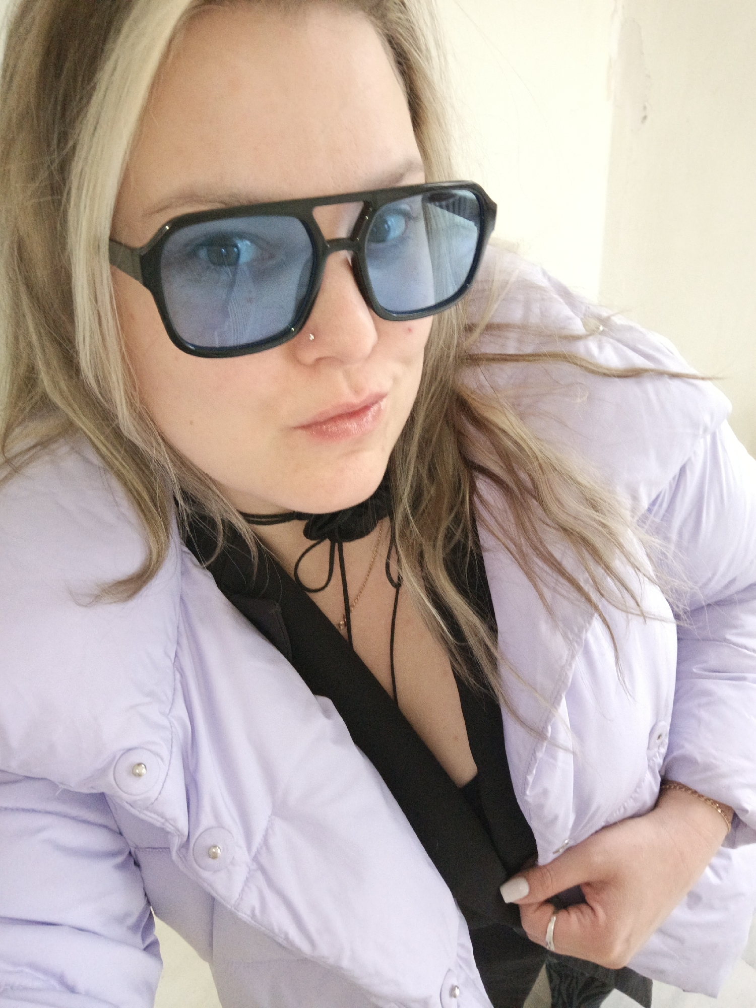 A woman is posing for a close-up selfie indoors, wearing blue-tinted square sunglasses and a light purple puffy jacket over a black top. A black choker necklace with dangling strings adorns her neck. She has blonde hair with darker roots, a nose piercing, and is wearing a ring on her right hand. The background appears to be a light-colored wall. There is no verifiable evidence to suggest that the location is Марковский, Russia.