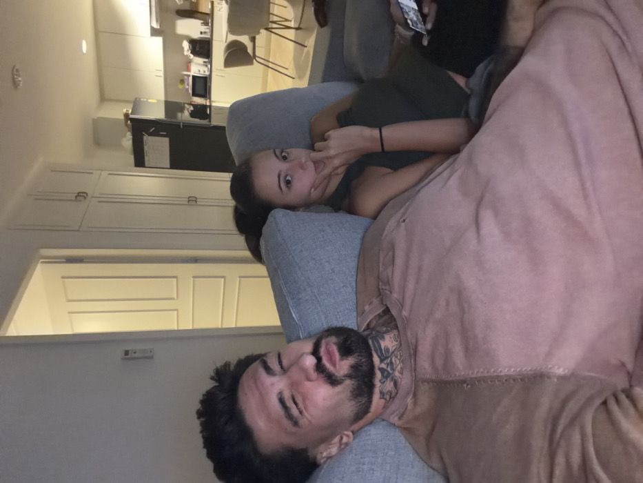 This image depicts an indoor scene in a modern apartment located in Bangkok, Thailand, with dim ambient lighting suggesting evening or night. Two individuals are seated on a light grey upholstered sofa.
On the left, a male subject, appearing to be in his 20s or 30s, is shown from the chest up. He has dark, short-to-medium length hair, a full beard, and a mustache. Distinctive tattoos are visible on his neck, featuring intricate patterns. He is wearing a light reddish-brown, long-sleeved shirt with visible seam details. His head is slightly tilted, and he is making a playful expression towards the camera with puckered lips and squinted eyes.
To the right of the male subject, a female subject, also appearing to be in her 20s or 30s, is seated on the same sofa. Her dark hair is pulled back, and she is wearing a dark green sleeveless top or dress. She is looking directly at the camera with a neutral expression, and her right hand is raised with the index finger lightly touching her chin. A portion of a white object, possibly a mobile phone or remote, is visible near her left leg or in her left hand.
The background reveals an open-plan living space. To the far left, a white wall with a decorative paneled door and a small wall-mounted control unit (possibly an air conditioner remote holder) are visible. To the right, an integrated kitchen area features white upper and lower cabinets, a dark-colored refrigerator, and a countertop with various small appliances including a microwave. Two light-colored chairs are partially visible, indicating a dining or breakfast nook. The overall environment is clean and furnished in a contemporary style.
