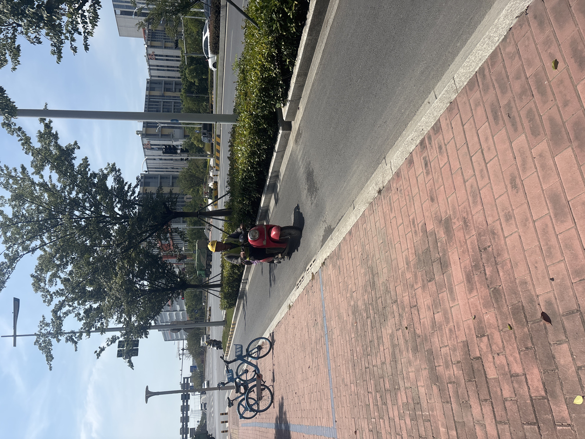 In Suzhou Industrial Park, China, a man wearing a yellow hard hat and a high-visibility vest rides a red scooter on a paved road. To his left, two blue bicycles are parked on a brick-paved pedestrian walkway. The scene is bordered by green bushes and trees, with modern buildings visible in the background under a clear blue sky with scattered clouds. Traffic lights and streetlights are also present along the road.