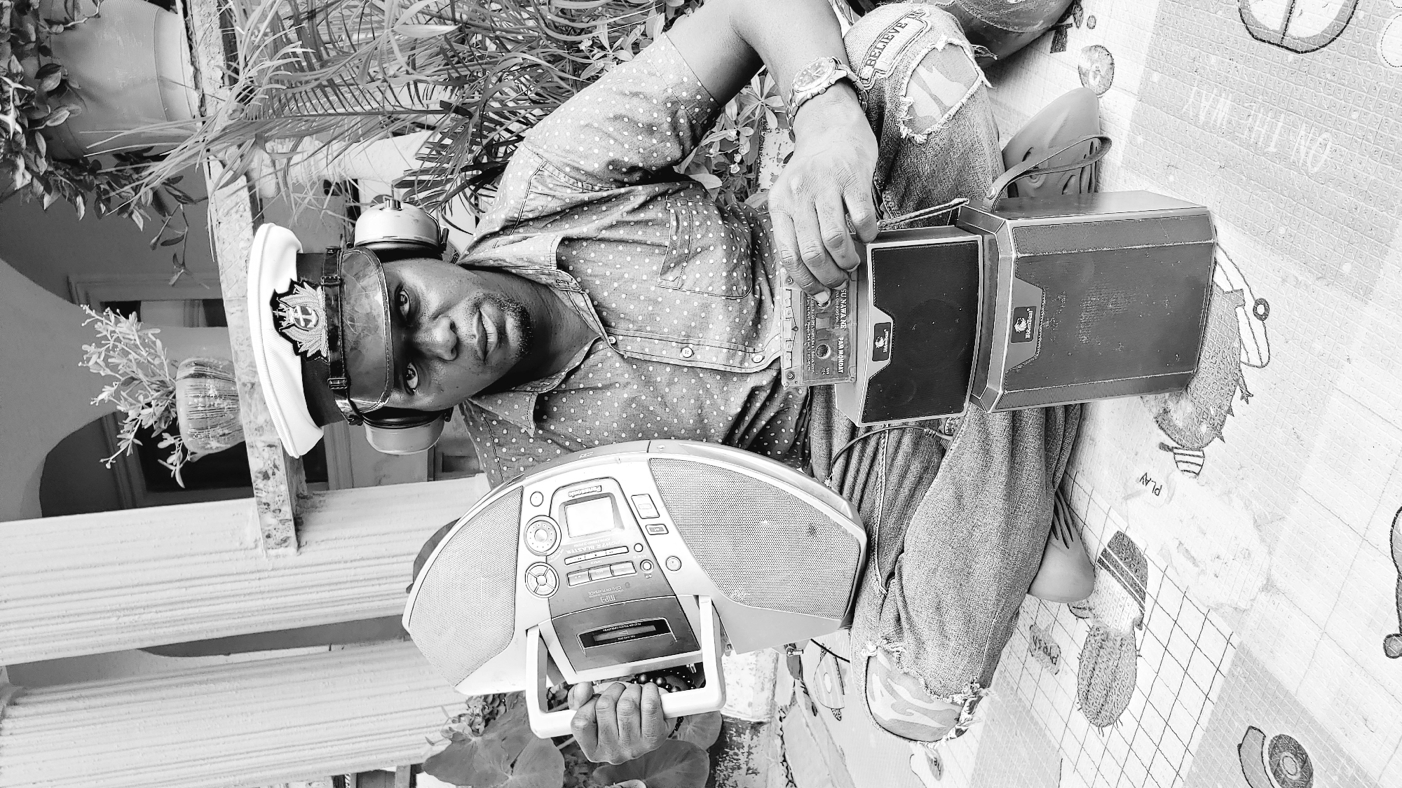 A black and white, eye-level, medium shot captures a Black man sitting cross-legged on a patterned mat outdoors. He is wearing a light-colored, polka-dotted short-sleeved shirt, ripped blue jeans, headphones, and a white captain's hat with a dark brim and emblem. The man is holding a large, light-colored portable CD player in his left hand and a cassette tape in his right hand, which he holds over a stacked pair of black speakers. He is looking directly at the camera with a serious expression. The background features architectural elements that suggest a building's facade, including pillars and an arched opening. Lush green foliage is visible to the right of the man. The setting appears to be a residential outdoor space.