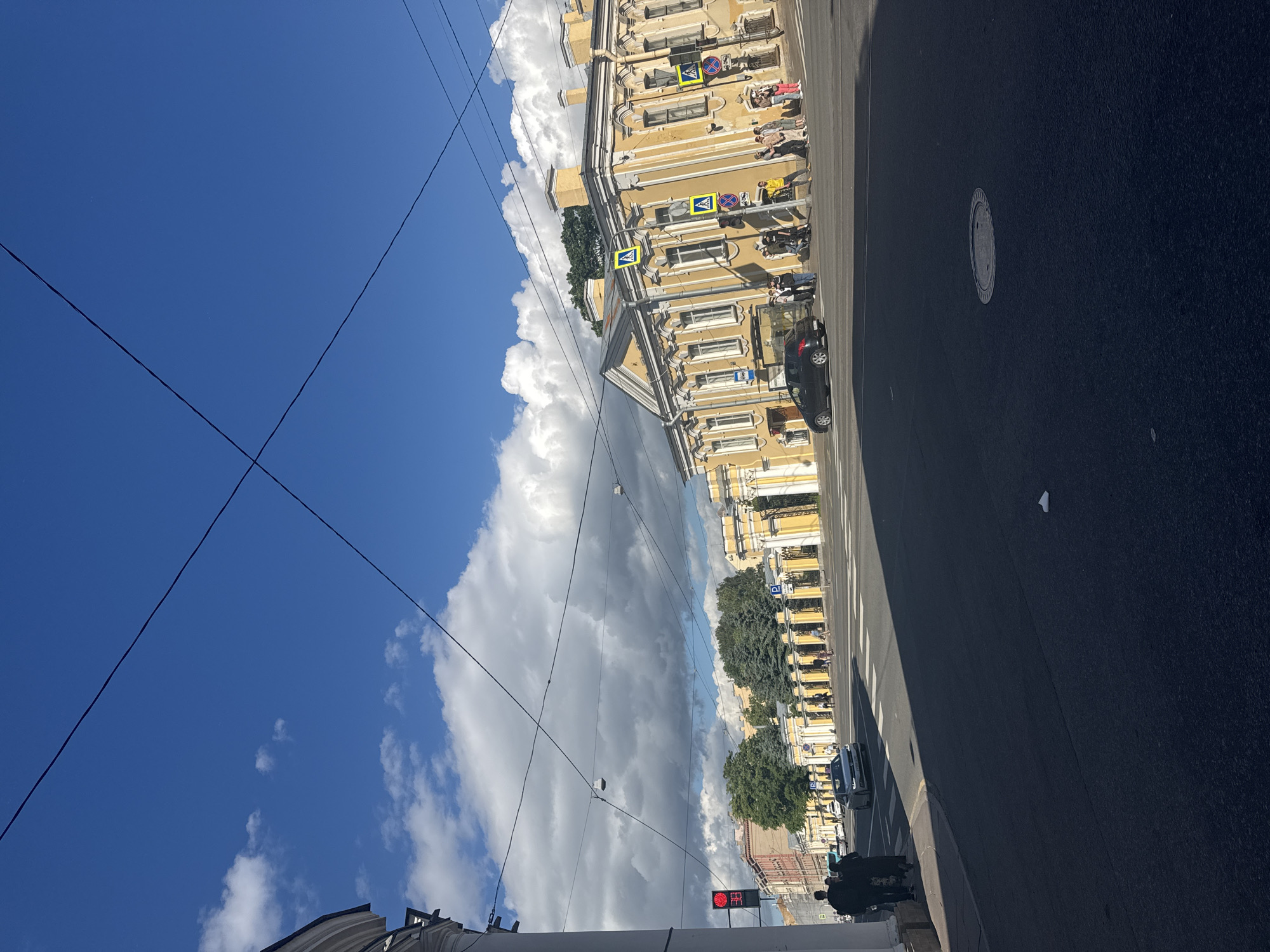 This is a rotated, eye-level, medium shot of a city street scene, likely in Saint Petersburg, Russia, during daytime. The sky is a vibrant blue with scattered, large white clouds. Power lines crisscross the sky, a common sight in urban areas.
The scene features a tall, yellow building with multiple windows, classic architectural details, and white trim. Some of the windows appear to be balconies or have awnings. Across the street from the building is a dark asphalt road, marked with white pedestrian crossing lines.
Several people are visible on the sidewalk in front of the building, some sitting on benches and others standing. A few cars are on the road, including a dark-colored sedan. A traffic light displays a red signal, indicating that traffic is stopped. A manhole cover is visible on the road. Trees line the sidewalk in front of the building, their green foliage contrasting with the yellow facade.
The lighting suggests it's a sunny day, casting sharp shadows. The overall mood appears to be calm and ordinary, depicting a typical urban street during daylight hours. No discernible text is visible in the image.