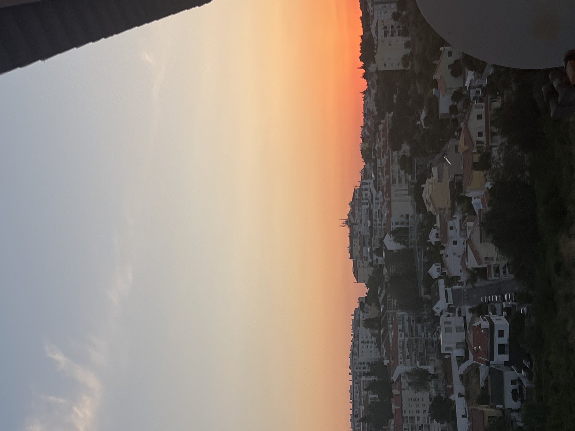 A high-angle, medium shot captures a residential area of Cascais, Portugal, at sunset. The sky transitions from pale blue at the top to a vibrant orange-red near the horizon, suggesting the sun is setting. A sliver of a dark structure, possibly a roof overhang, is visible in the top right corner. Below the sky, a dense collection of buildings with varying architectural styles, mostly white and beige with red-tiled roofs, covers the landscape. These buildings range from multi-story apartment blocks to smaller detached houses. Trees and greenery are interspersed throughout the urban setting, some appearing as dark silhouettes against the illuminated sky. A road with parked cars can be seen winding through the neighborhood. In the foreground, on the right side, a portion of a large, light-colored satellite dish is visible. The overall impression is of a quiet, developed urban neighborhood bathed in the warm glow of twilight.