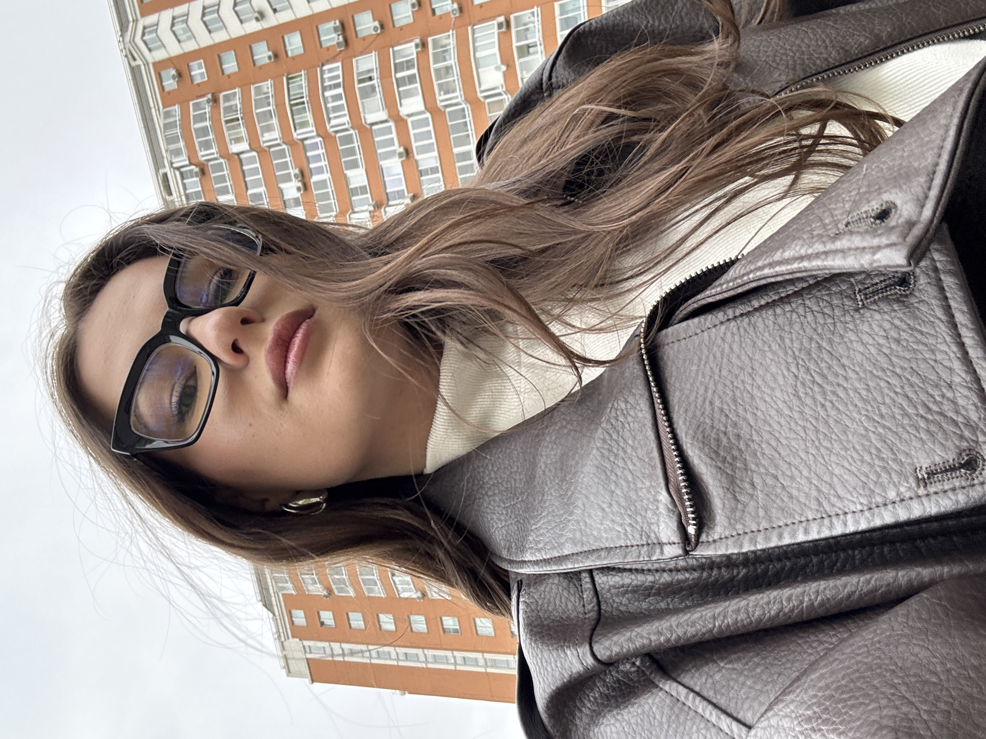 A low-angle, slightly off-kilter close-up photograph shows a young woman with long brown hair, wearing black-framed glasses, a white turtleneck sweater, and a dark leather jacket. The jacket is partially zipped, revealing the sweater underneath. The woman's face is angled towards the camera, with her gaze directed slightly downwards. In the background, a tall, multi-story building with a red and white facade is visible against a gray, overcast sky. The building appears to be located in an urban area, likely Vnukovo, Russia.
