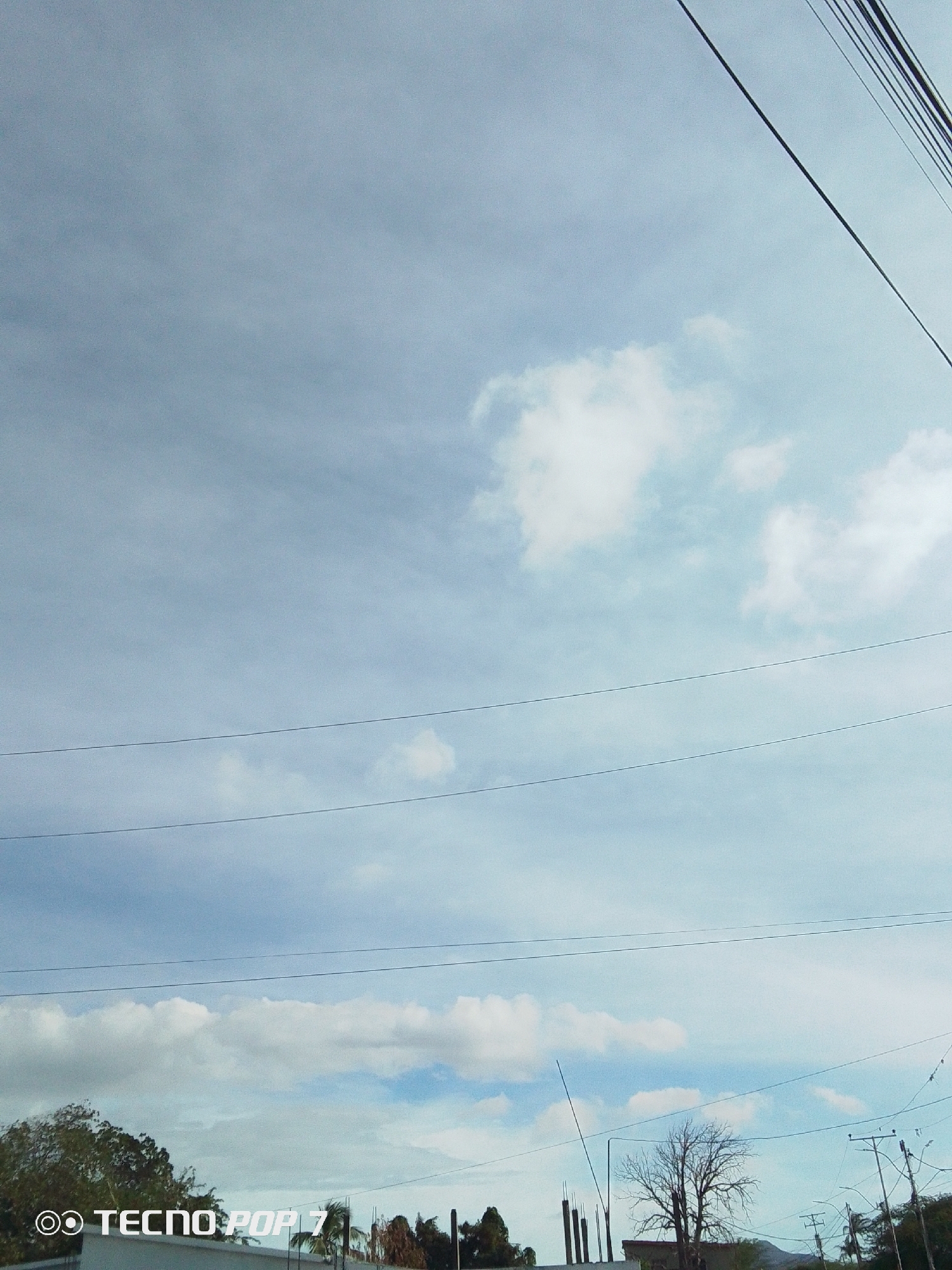 The image shows a cloudy sky with several power lines running across the frame. In the lower part of the image, there are trees, vegetation, and buildings with a few unfinished constructions. The image appears to have been taken in a residential area, with electrical posts visible. The location is Altagracia, Venezuela. There are no people visible in the frame.