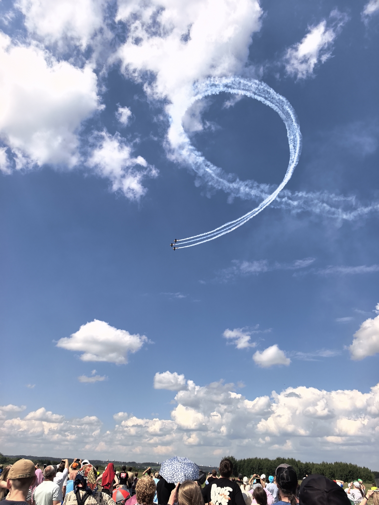 An aerial display is taking place under a bright blue sky with scattered white clouds. Three small, dark planes fly in formation, trailing white smoke that forms a large, incomplete circle in the sky. The planes are positioned in the upper right quadrant of the image. The foreground shows a crowd of people gathered, facing away from the camera and towards the sky, presumably to watch the air show. They are standing in a field with a line of trees visible in the distance.