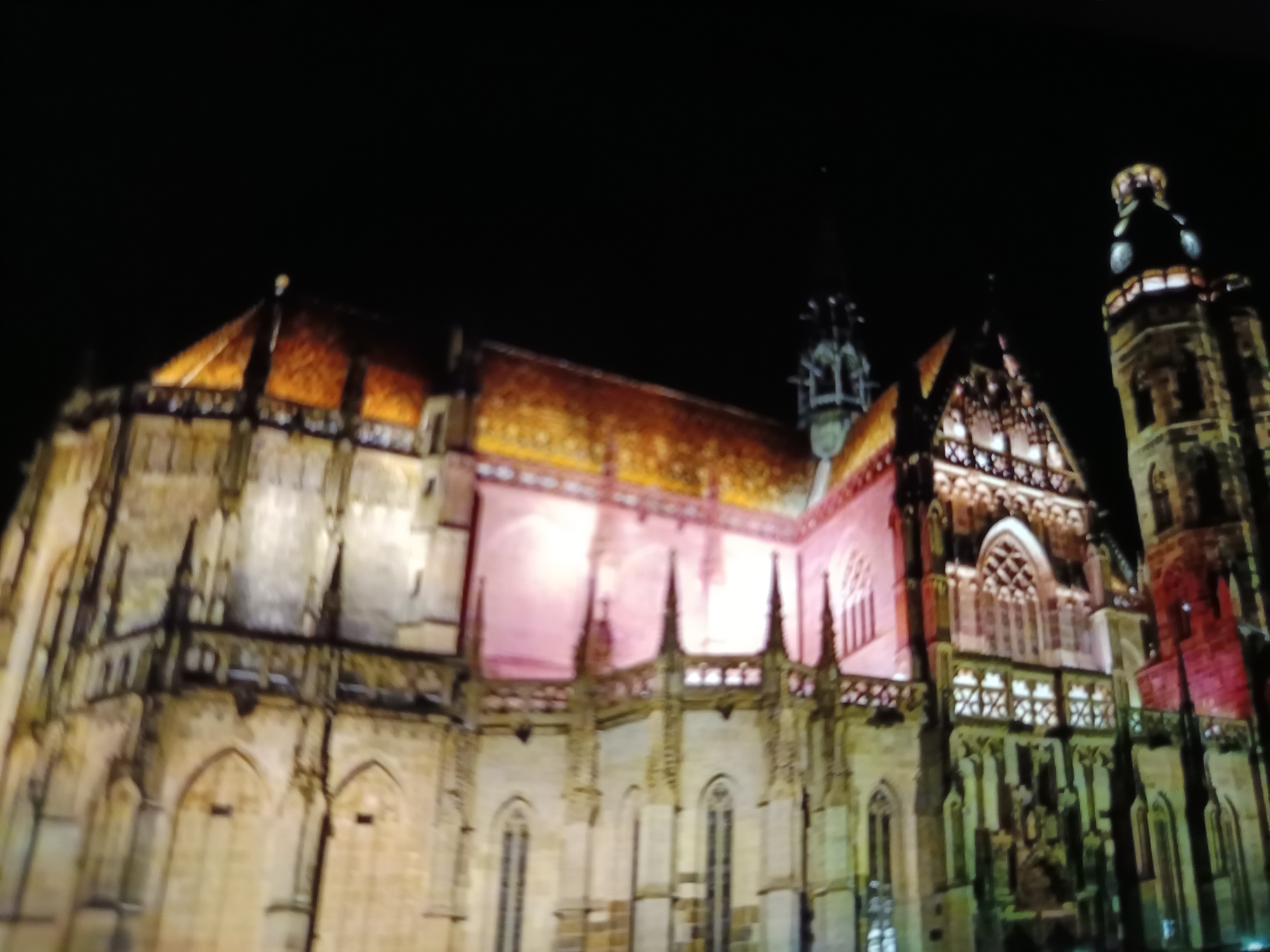 The image depicts a night view of St. Elisabeth Cathedral in Košice, Slovakia, illuminated by artificial lights. The cathedral is a Gothic masterpiece, characterized by its impressive architecture, including soaring spires, intricate stonework, and a distinctive tiled roof. The lighting casts a warm glow on the facade, highlighting the details of the structure and creating a dramatic effect against the dark night sky. The scene is devoid of people, focusing solely on the grandeur of the cathedral and its surroundings, which appear to be dark and possibly deserted at this hour. The overall impression is one of solemn beauty and historical significance.