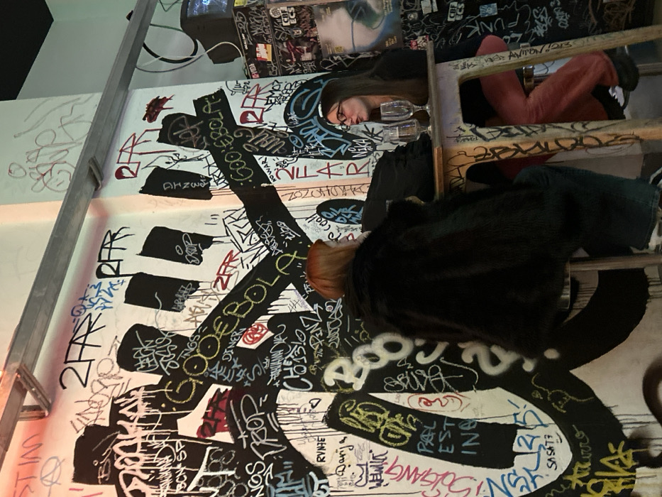 A cluttered composition features two individuals sitting on a ladder against a graffiti-covered wall. The wall is adorned with dense layers of writing, symbols, and illustrations in black and various other colors. One person is dressed in dark clothing and has auburn hair, while the other is wearing a dark top and pink bottoms. Two wine glasses are placed near them. The ladder is positioned against the wall, adding to the overall chaotic aesthetic.