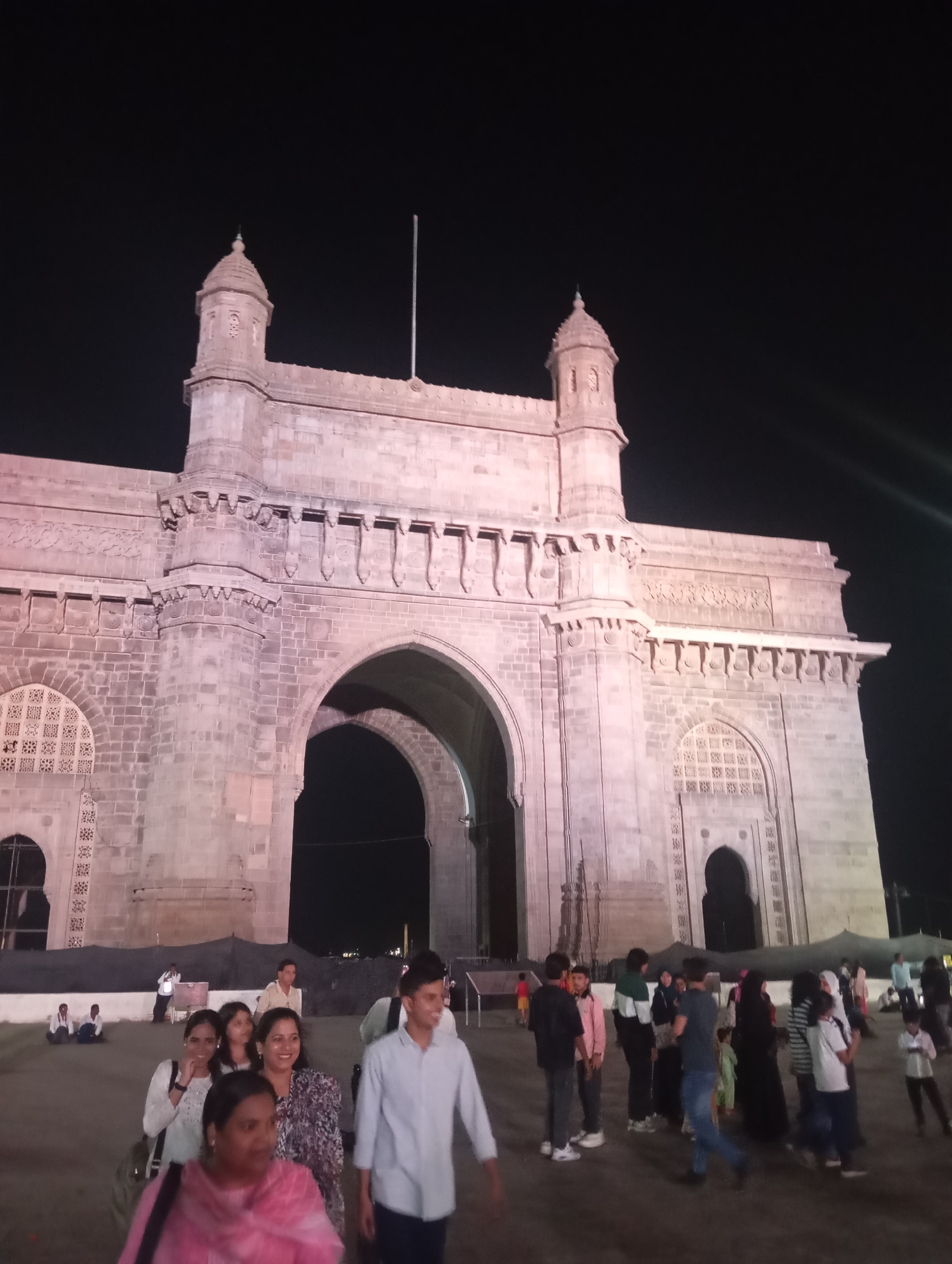 The image depicts the Gateway of India in Mumbai, India at night. The large arch monument, illuminated with artificial lighting, dominates the scene. A crowd of people is present in the foreground, including groups standing and conversing, and others walking. The background is a dark sky.