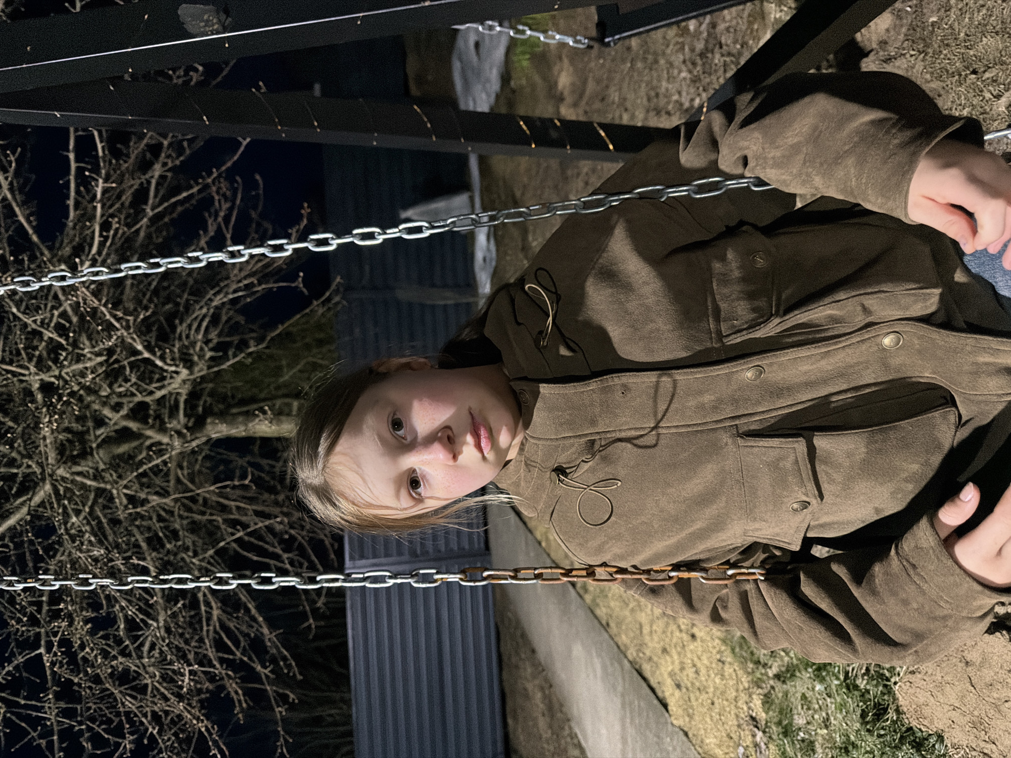 A photograph shows a juvenile female seated on a swing. She is wearing a brown corduroy jacket. The swing is suspended by metal chains attached to a dark metal frame. Behind her is a metal corrugated fence, bare tree branches, and patches of grass. The photograph appears to be taken outdoors at night. The exact location in the city of Dubovaya, Russia cannot be determined from this image.