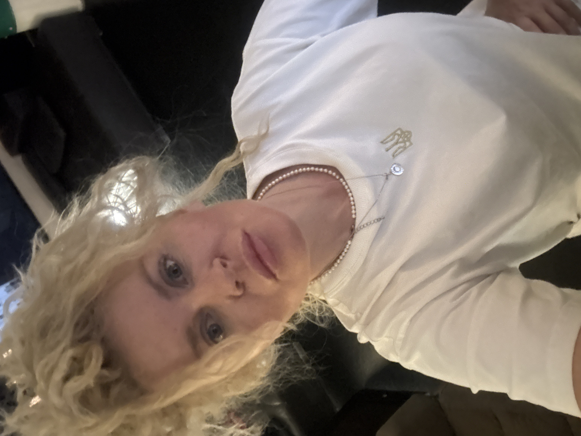 A woman with blonde, curly hair is seen from the chest up, looking directly at the camera. She is wearing a white t-shirt and a pearl necklace with a pendant. The background is dark and appears to be indoors, with some blurred objects that suggest furniture or a room setting. The lighting is somewhat dim, with a bright light source coming from the top left, illuminating the woman's face and hair. There are no clear indications of her location within Nanhai District, China.