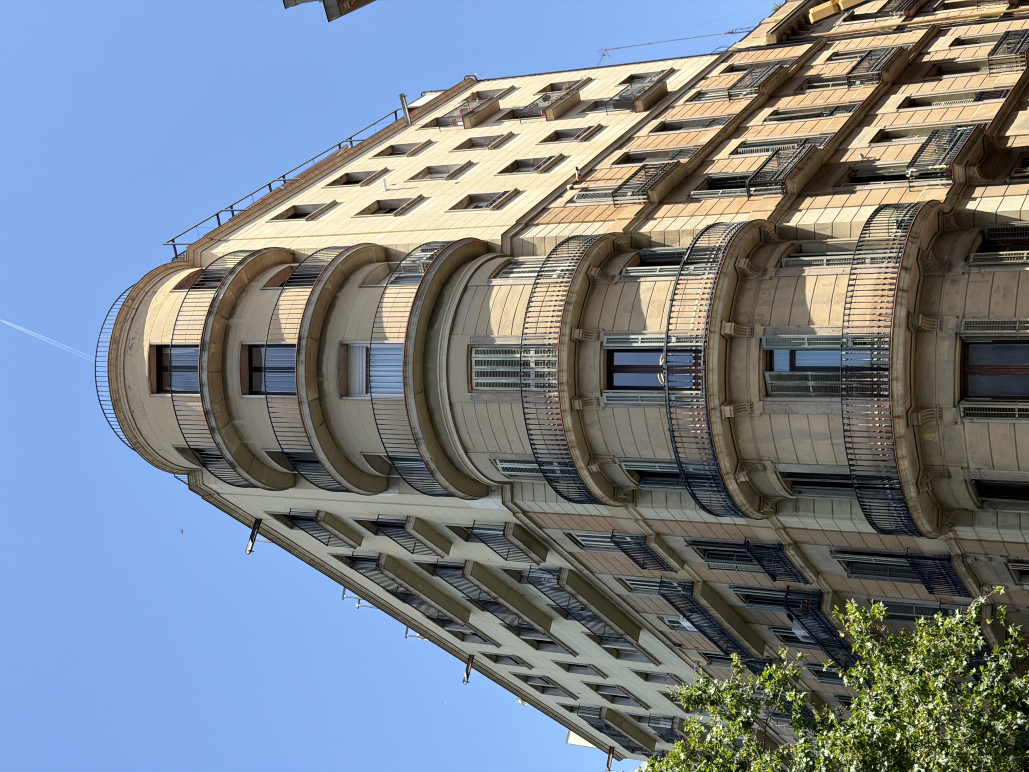 The image shows a multi-story building in Barcelona, Spain, with a light tan facade and a curved corner section. The building has multiple balconies with black iron railings on each floor, and many of the windows have green shutters, some of which are open. The sky above is clear and blue, with a faint contrail visible from an airplane. A large green tree with dense foliage is visible on the left side of the building. The architecture suggests an older European style, common in cities like Barcelona. No people are visible in the image. The overall scene is one of urban architecture under a clear sky.