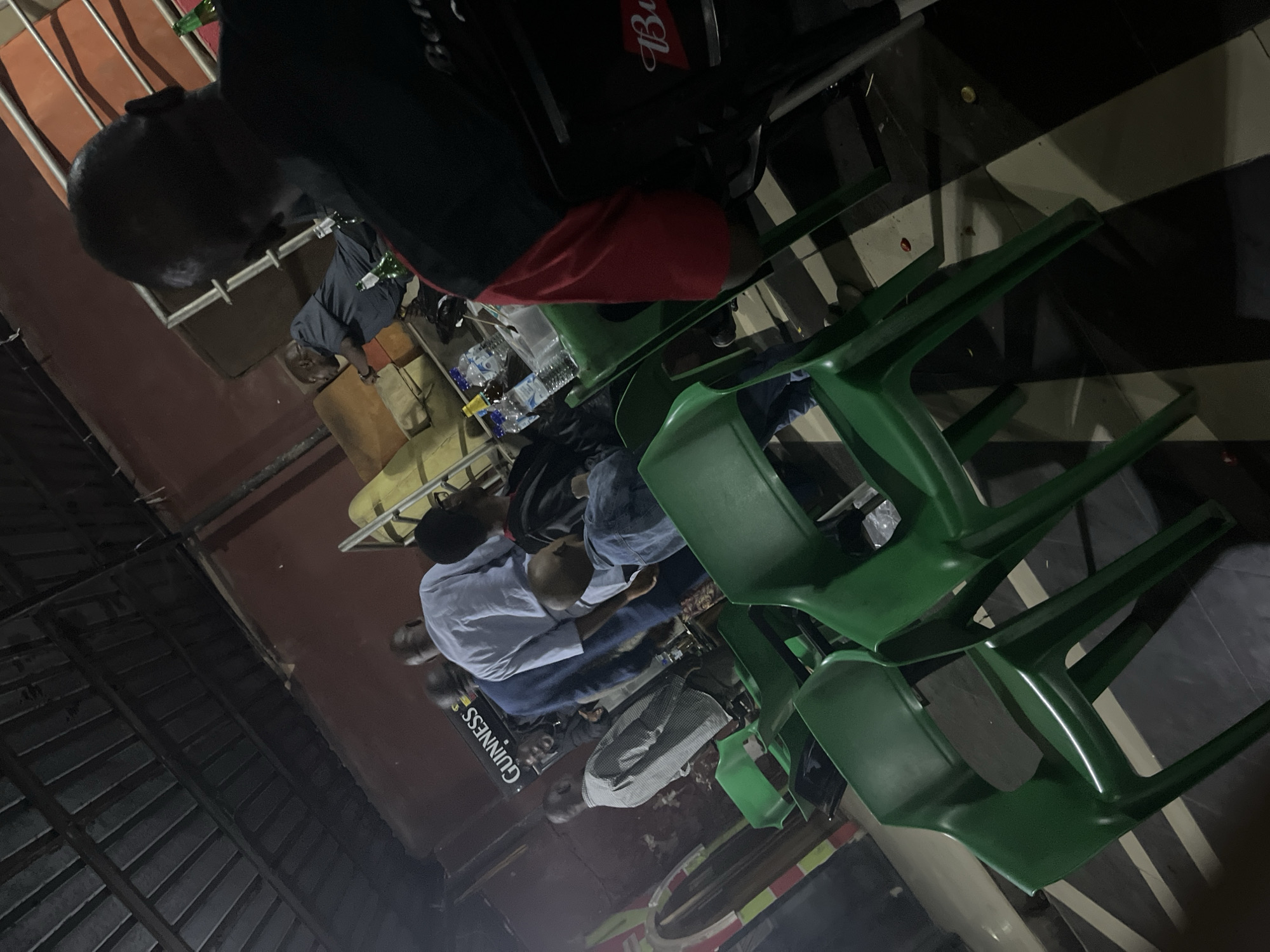 The image depicts a dimly lit outdoor gathering, possibly a bar or a casual seating area, with several individuals present. The scene appears to be in the city of Jos, Nigeria.
In the foreground, several bright green plastic chairs are arranged, some of which are empty, while others appear to have people sitting in them or nearby. The chairs are a prominent visual element, suggesting a functional, perhaps informal, setup for patrons.
Behind the chairs, a group of men are seen mingling. One man in a light blue shirt is standing, facing away from the camera. To his left, another man in a checkered shirt is also standing and appears to be engaged in conversation or observing something. Further to the left, a prominent "Guinness" logo is visible on a dark background, indicating a possible sponsorship or product placement.
In the midground, a person in a dark denim jacket is seated, holding what appears to be a child on their lap. This interaction adds a domestic or family element to the scene, contrasting with the more social atmosphere suggested by the other individuals.
To the right of the frame, a man in a black shirt with red sleeves is the most prominent figure. He is seated and facing away from the camera, his back and the side of his head visible. He appears to be part of a group, with other individuals partially visible around him. Bottles, possibly containing drinks, and other containers are present on a table in front of this group.
The environment is an outdoor space with a corrugated metal roof structure overhead, supported by metal poles. The walls appear to be painted in a reddish-brown hue. The ground has a checkered pattern of dark and light colored tiles or surfaces. The lighting is artificial and casts shadows, contributing to the low-light ambiance of the photograph. The overall impression is of a relaxed, social gathering in a casual setting.