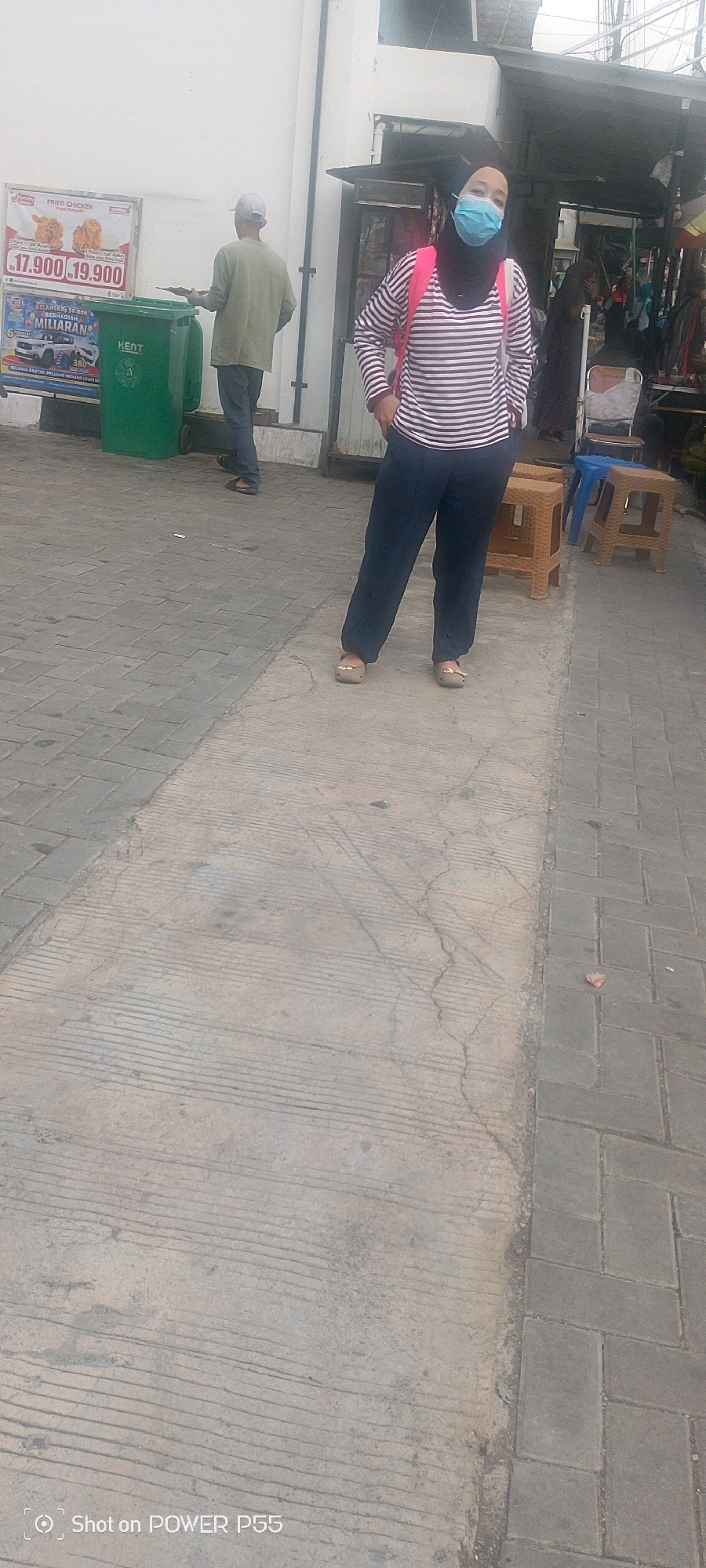 The image depicts an outdoor scene on a paved surface. In the foreground, a woman wearing a blue face mask, a black hijab, a striped long-sleeved shirt, and dark pants stands with her hands in her pockets. She has a pink backpack slung over her shoulders. To her left, a man wearing a light-colored cap, a long-sleeved shirt, and dark pants stands holding a flat object. Behind them, there are green trash bins and a sign that reads "FRIED CHICKEN" and pricing information. Further in the background, there are stalls and various people. The environment suggests a marketplace or outdoor commercial area. The ground is a mix of concrete and interlocking pavers. A watermark at the bottom reads "Shot on POWER P55".