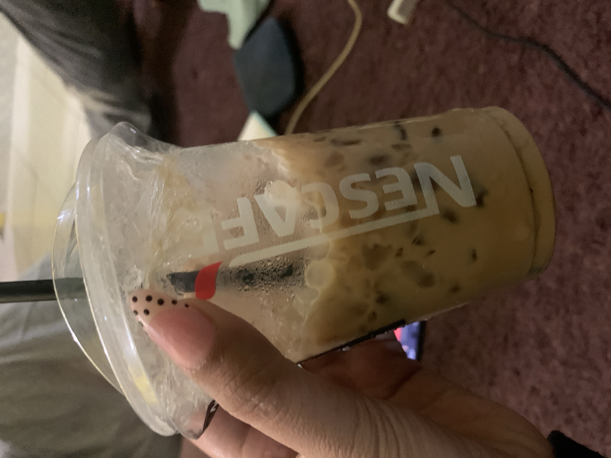 A person's hand, with light pink, almond-shaped nails decorated with black polka dots, holds a clear plastic cup filled with iced coffee. The cup has condensation on the outside and the Nescafé logo is visible on the side in white lettering. A black straw with a red accent is also present. The background shows a blurry, dark red carpeted surface and what appears to be a person's leg in dark pants. The scene suggests an indoor setting, possibly a living room or lounge area. There is no information in the image that indicates a specific location in Jajag, Indonesia.