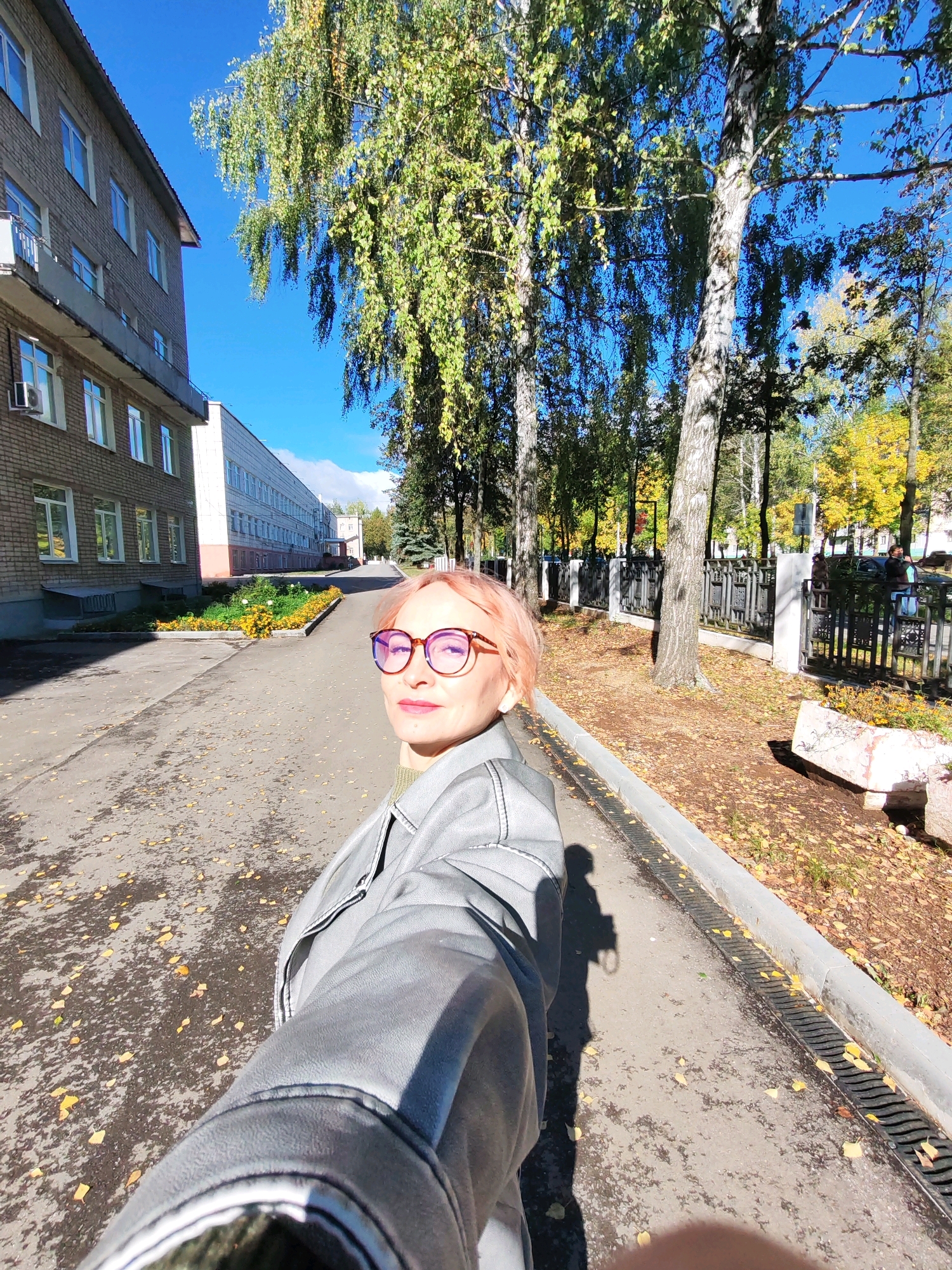 A woman with short, light pink hair and round, red-tinted glasses is taking a selfie outdoors. She is wearing a light gray jacket over a green top. The background shows a paved walkway lined with trees and a multi-story brick building on the left. The ground is covered with fallen yellow leaves, indicating an autumn setting. The sky is clear and blue. The image appears to be taken in a park or on a street in the city of Chaikovsky, Russia, likely near an educational institution or administrative building, as suggested by the architecture and manicured flower beds.