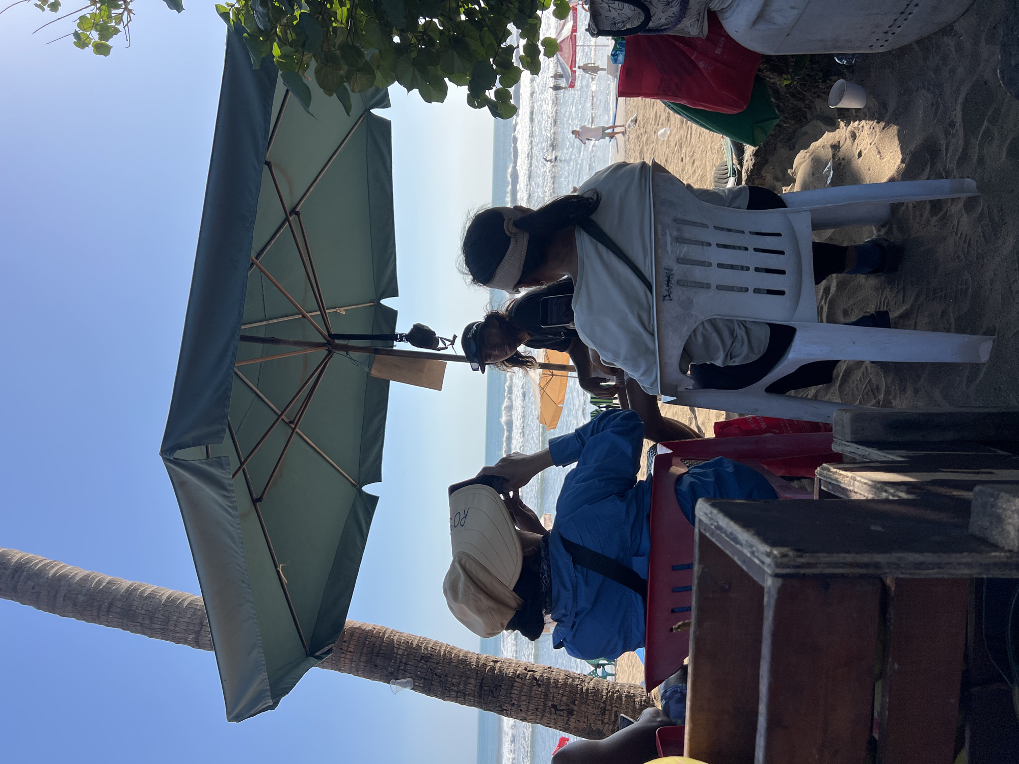 The image depicts a beach scene in Kuta, Indonesia, likely at mid-day given the bright sunlight and clear sky. In the foreground, three individuals are seated on chairs under a large green umbrella. The person on the left, facing away from the camera, wears a blue jacket and a wide-brimmed light-colored hat. They are holding a phone up to their face, seemingly taking a photo or video. To their right, a person with dark hair, wearing a dark shirt, is also looking forward, holding a phone. The third person, on the right, is also facing away from the camera, wearing a light-colored long-sleeved shirt and a headband. They are seated in a white plastic chair. The chairs are placed on sandy ground.
Behind the seated individuals, the ocean is visible with moderate waves and several people in the water, some appearing to be surfers. Other beach umbrellas and chairs are scattered on the sand. A palm tree trunk is prominent on the left side of the frame. Various objects are present around the seating area, including a wooden crate in the lower left corner, red and green bags, and a white cooler. The overall impression is a relaxed beach day, possibly at a cafe or seating area overlooking the ocean.