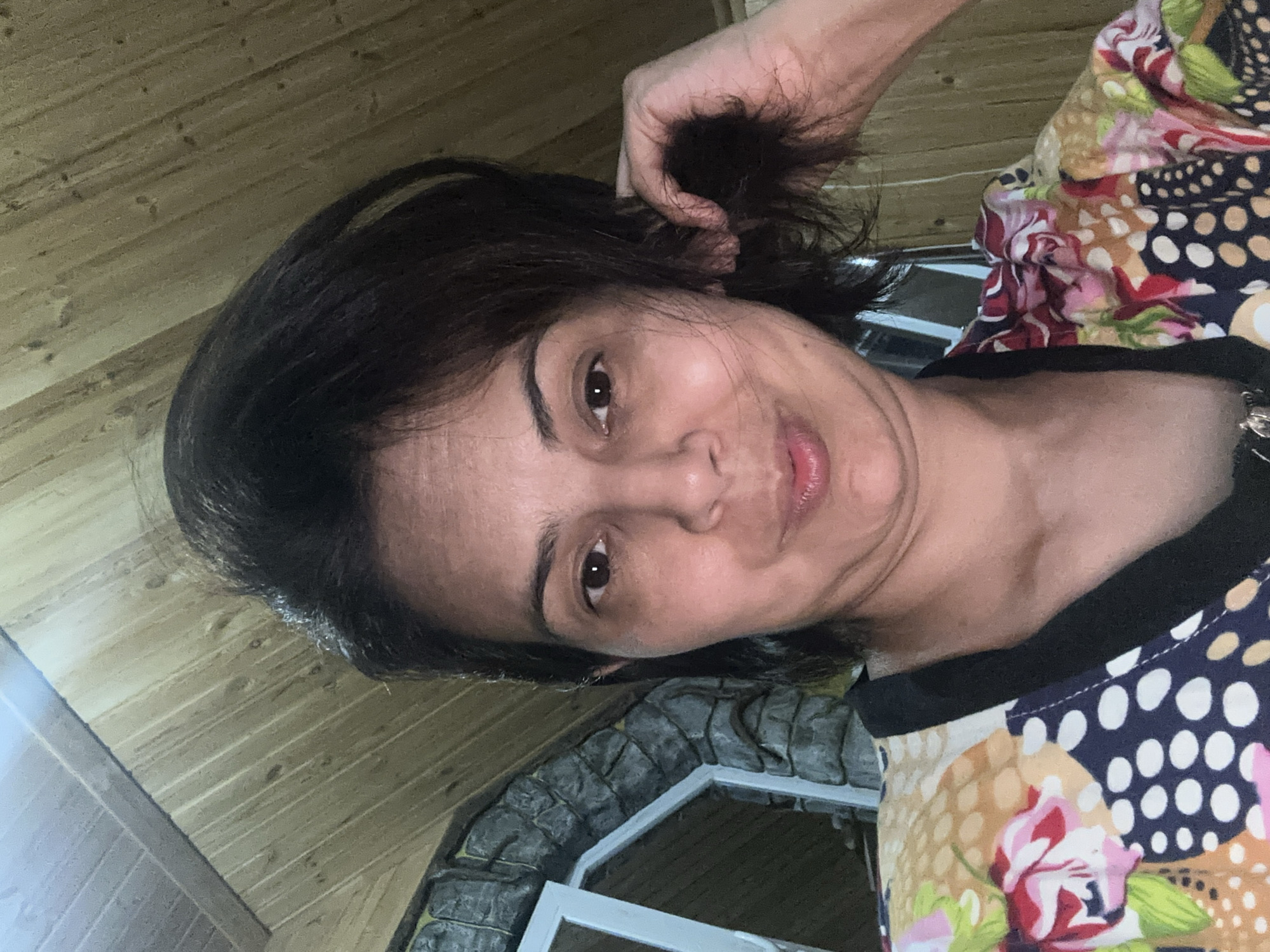 A woman is taking a selfie in what appears to be a room with a wooden ceiling and stone walls. The woman has dark brown hair and is wearing a colorful patterned shirt. She is looking at the camera and holding a strand of her hair with her right hand. The background includes part of a window and stone masonry. The lighting is dim, suggesting an indoor or evening setting.