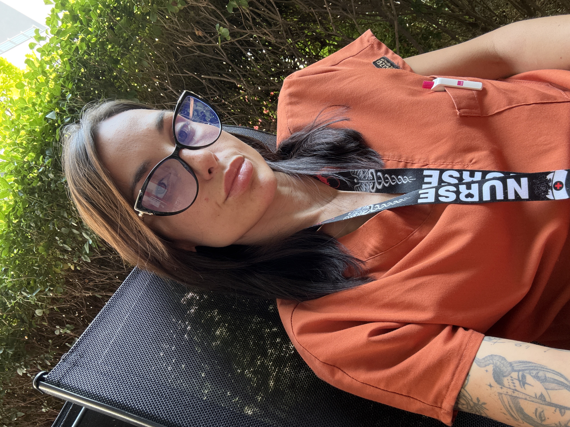 In Pleven, Bulgaria, a person wearing an orange scrub top and distinctive cat-eye glasses is seated in a black mesh chair, with lush green foliage forming the background. The individual has dark hair, some tattoos visible on their left arm, and a lanyard with "NURSE" printed on it hanging around their neck. A pen is tucked into the chest pocket of the scrub top. The lighting suggests it is daytime, with shadows indicating a sunny environment.