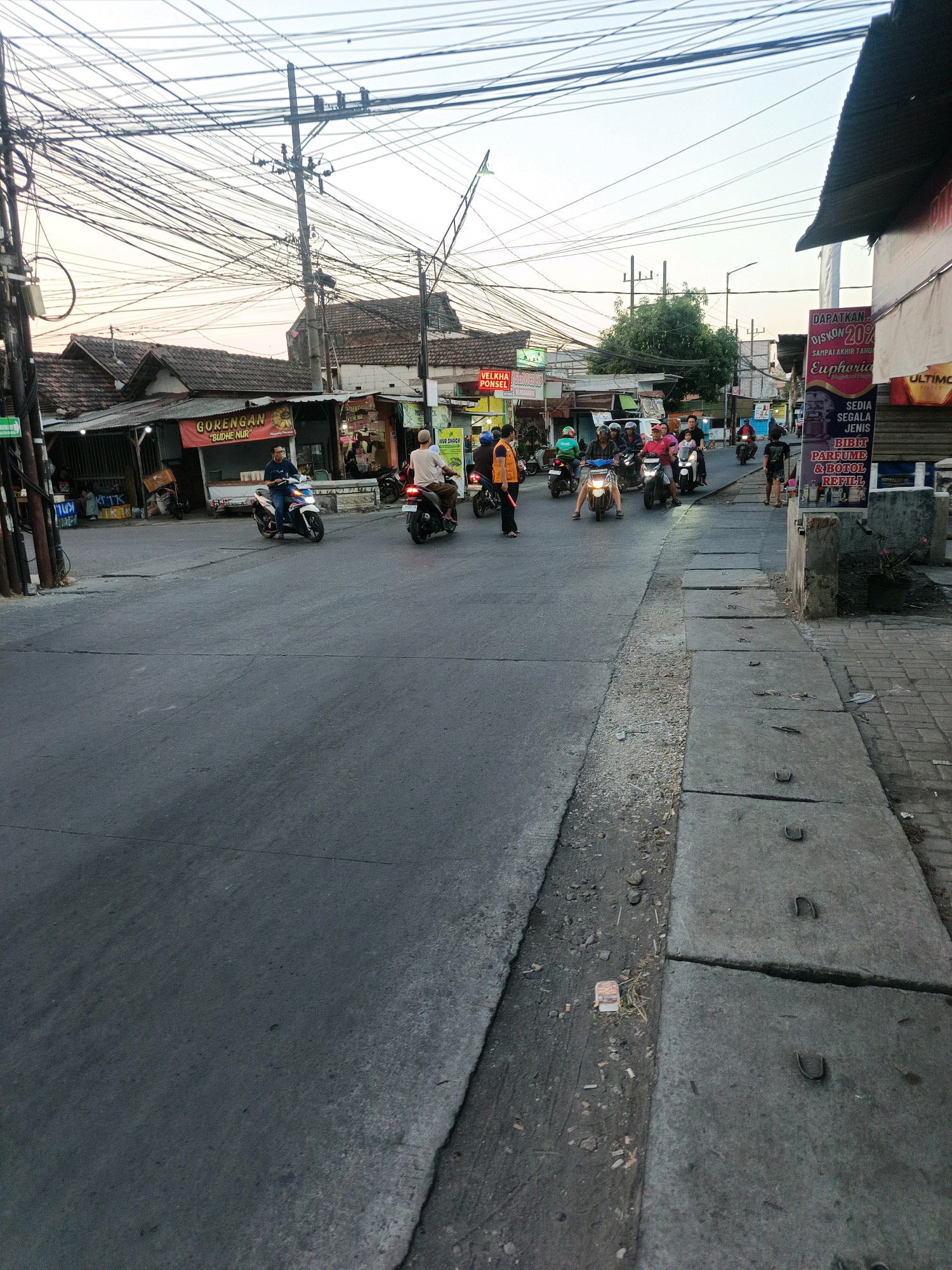 The image depicts a street scene in Kwangsan, Indonesia, characterized by a dense network of overhead electrical wires. The road surface is asphalt, with a concrete sidewalk on the right side containing several manhole covers. Several motorcycles are visible on the road, with riders navigating the traffic. To the left, there are small shops and buildings with tiled roofs. One shop has a red banner with white text that appears to be an advertisement. A man in an orange vest and holding a red baton stands on the side of the road, seemingly directing traffic or in a similar role. The sky is overcast, suggesting it might be late afternoon or early morning. The overall impression is one of a busy, functional urban environment.