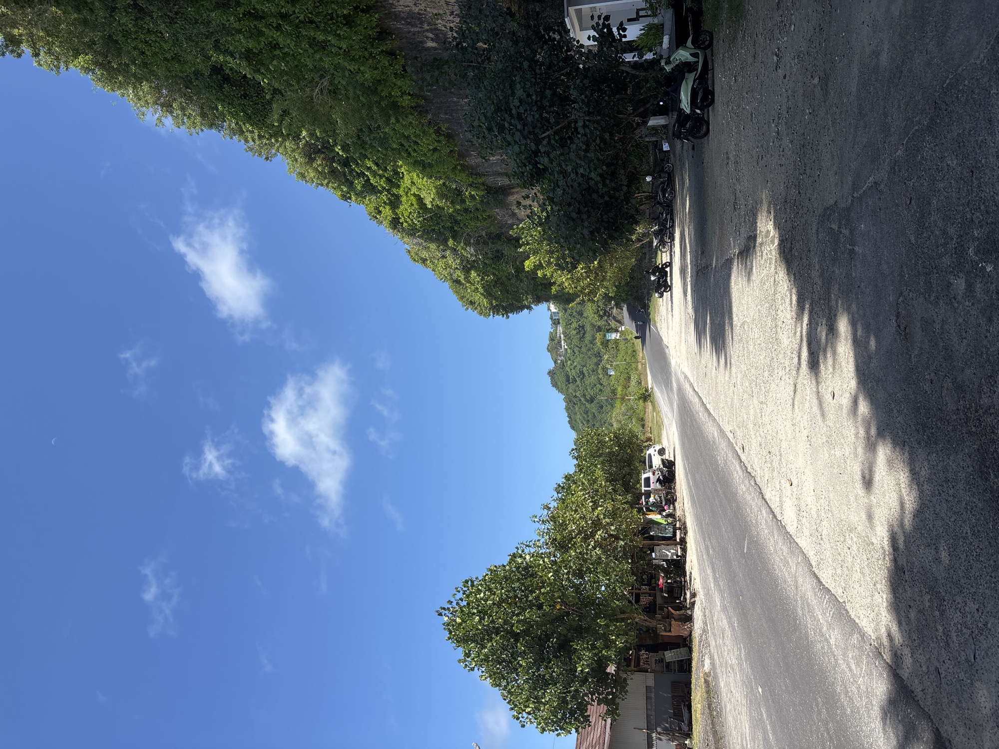 The image captures a road in Kutuh, Indonesia. The road runs diagonally across the frame, flanked on both sides by lush green vegetation and trees. On the right side of the road, several parked motorbikes are visible near a building, casting shadows on the road surface. The opposite side of the road also features dense vegetation. The sky above is a clear blue with scattered white clouds, and a sliver of the moon is visible in the upper left corner.