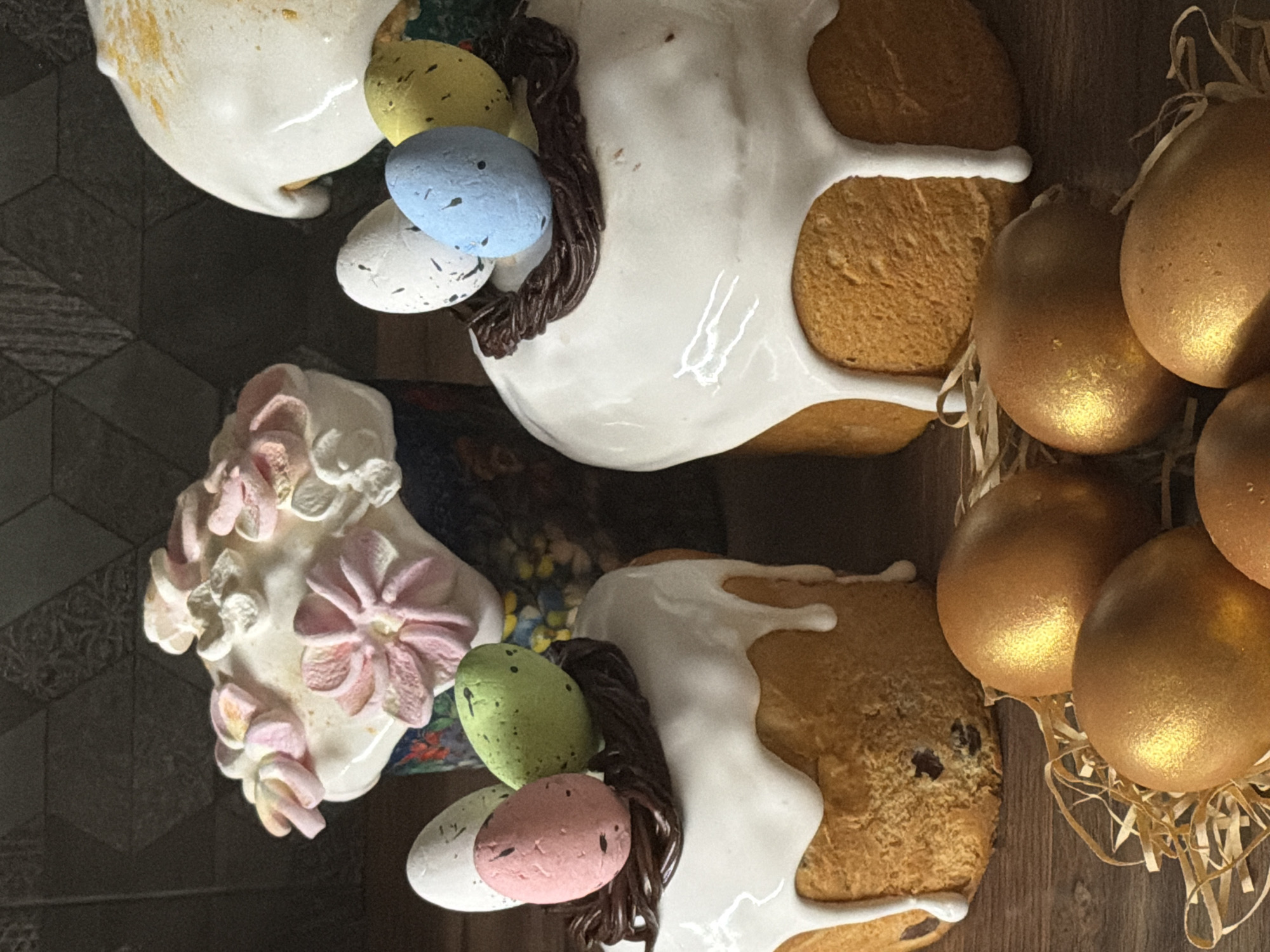 The image depicts a scene likely related to Easter celebrations, captured indoors. It features two Easter cakes, each decorated with white icing and a nest of colored eggs. One cake has white and pink flower decorations. Adjacent to the cakes are golden eggs arranged in a straw nest. The image is a reflection, possibly in a mirror or glass surface. Given that the image origin is Perm, Russia, the scene is likely located within that city.