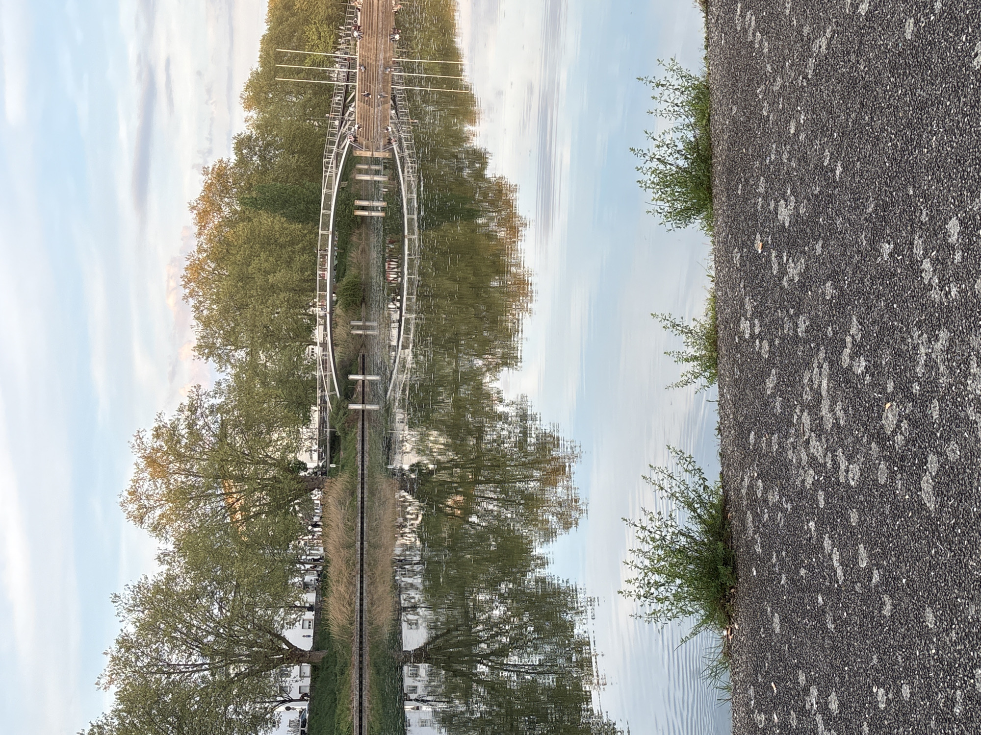 The image shows a canal with a mirrored reflection of the environment above the canal. There is a pedestrian bridge over the canal, with several people on it. Trees are visible on either side of the canal, as well as buildings. The right side of the image features a paved walkway with some bushes growing along the edge of the canal. The image seems to be taken somewhere in Strasbourg, France.