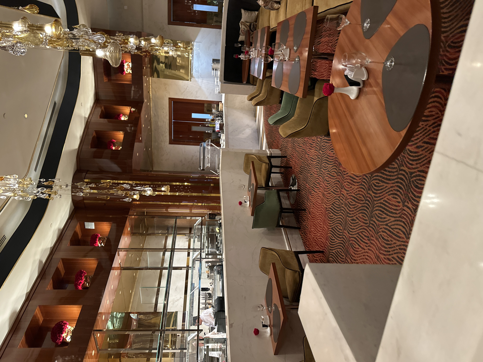 This image captures the interior of a luxurious restaurant or upscale dining area. The scene features multiple tables, some round and some rectangular, with polished wooden tops, dark grey placemats, wine glasses, and small white vases each holding a single red rose. The chairs are upholstered in varied tones of olive green and light brown/beige, arranged around the tables. The floor of the main dining space is covered with a distinctive patterned carpet in orange/reddish hues with black, wavy lines.
To the left, a long, dark wooden wall is adorned with several recessed alcoves, each housing a vibrant red floral arrangement, possibly hydrangeas. Ornate, multi-tiered chandeliers made of glass and gold elements hang from the ceiling, providing warm illumination throughout the room. In the background, a serving area or open kitchen pass-through is visible behind glass partitions, with several staff members in white uniforms working behind a counter that displays kitchen equipment and food items.
The decor includes elements of dark wood paneling, white marble surfaces, and reflective glass, contributing to an opulent and refined atmosphere. The lighting suggests it is either evening or an indoor setting where artificial light is predominant. No specific text is visible within the frame. The overall impression is one of elegance and high-end hospitality in Estosadok, Russia.