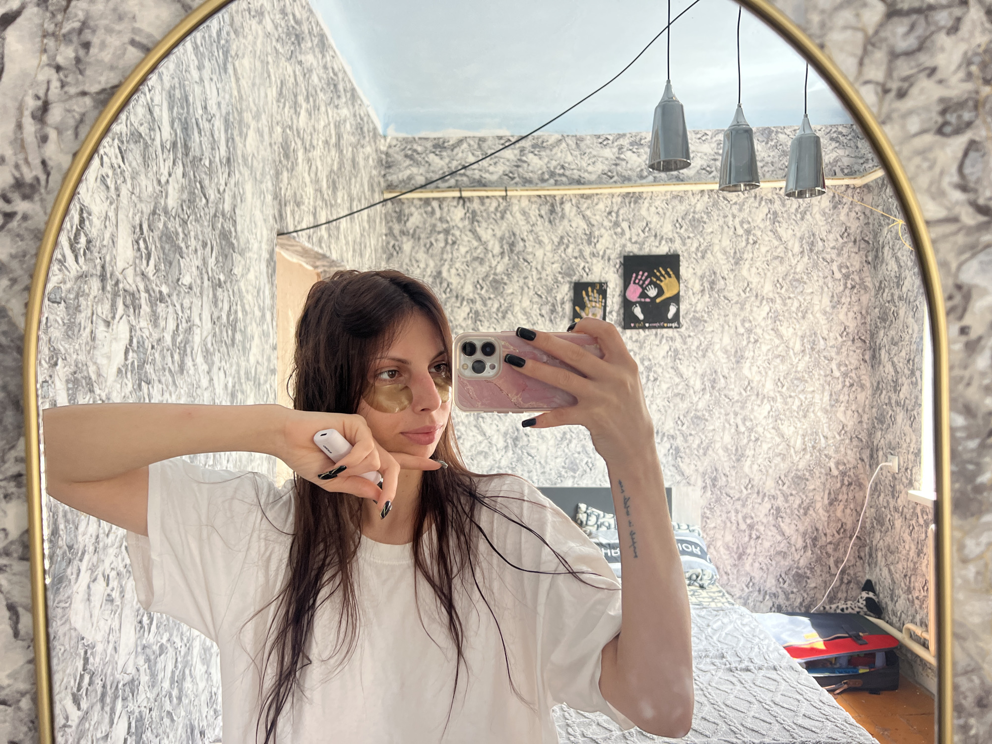 A young woman with long, dark brown hair, wearing a white t-shirt and gold under-eye patches, is taking a mirror selfie in a bedroom. She is holding a pink phone in her right hand and a white electronic device in her left. The room features textured gray wallpaper, a light blue ceiling, and three pendant lights hanging from the ceiling. A bed with gray bedding is visible in the background, along with some framed artwork on the wall. The mirror has a gold frame and is mounted on the wall. The scene is set in a residential interior.