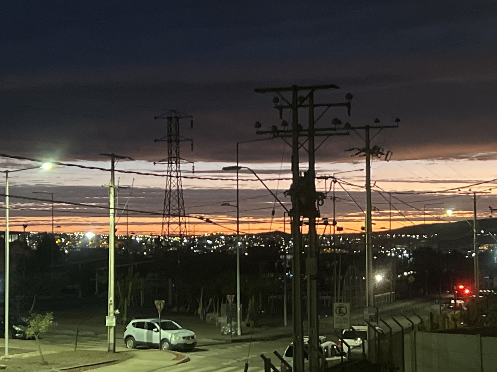The image depicts a twilight cityscape at dusk, with the sky exhibiting hues of dark blue and orange from the setting sun. In the foreground, several utility poles with electrical wires crisscross the frame, some carrying insulators. A large transmission tower is visible on the left side of the image, and another one is partially obscured on the right. A white car is parked on the right side of a paved road in the mid-ground, with another vehicle visible further down the road. In the background, a sprawling city lights up the horizon, with hills or mountains forming the distant backdrop. The scene appears to be an urban or suburban area, possibly on the outskirts of the city of Arica, Chile, given the presence of the transmission towers and the coastal proximity suggested by the vast expanse of lights. The overall impression is one of a developing urban landscape transitioning from daylight to night.