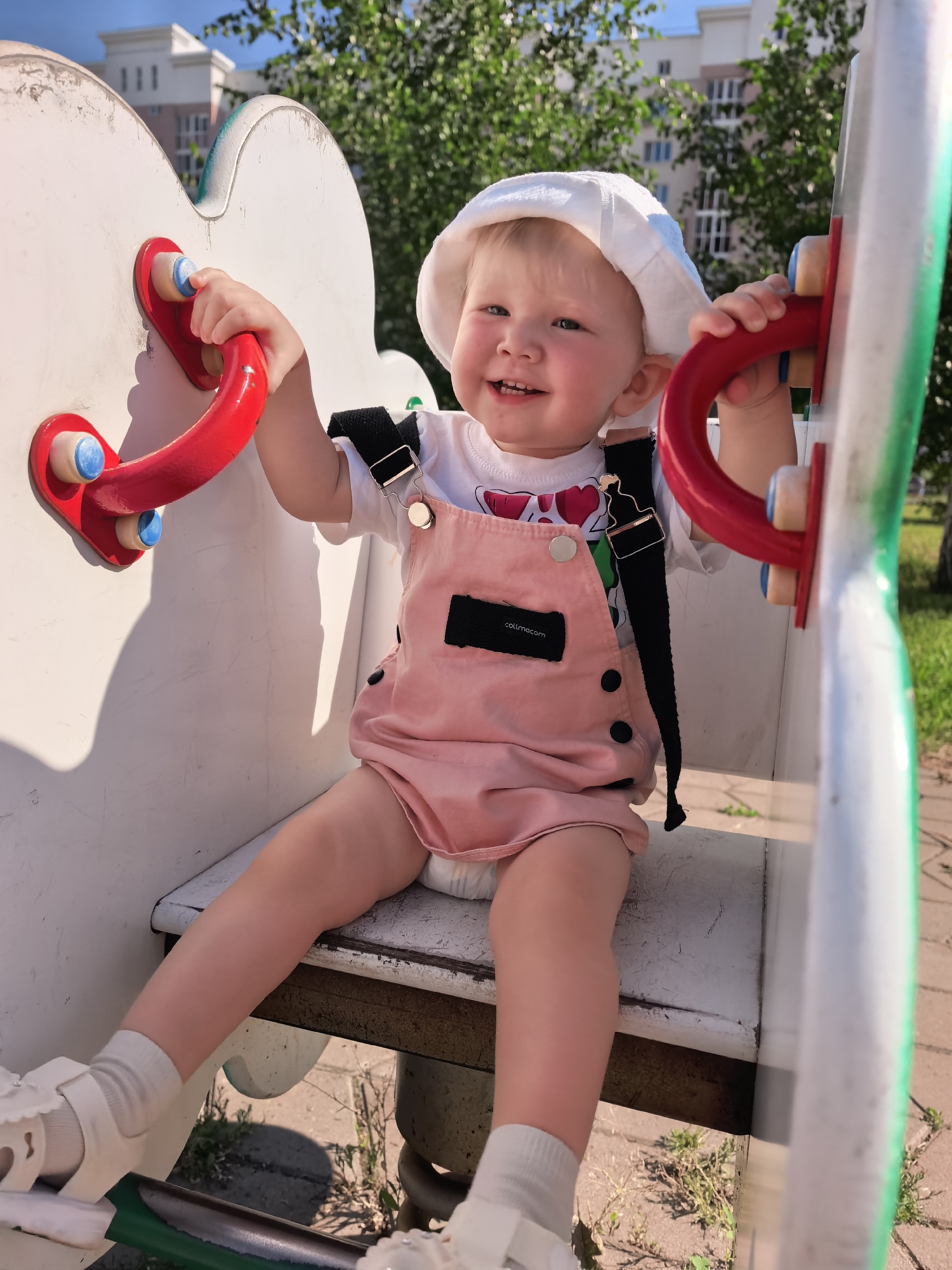A young Caucasian child, approximately 1-2 years old, sits on a white playground structure with red handles. The child wears a white bucket hat, a white t-shirt with a pink heart design, and a pale pink romper with black straps. They are smiling and looking at the camera. The playground structure is white with some green and white accents on the right. In the background, there are green trees and a modern building with a white facade and several floors, typical of apartment buildings in Sterlitamak, Russia. The ground beneath the playground is paved with light-colored square tiles and scattered with dry grass. The overall scene suggests a sunny day at a local park or playground in the city.