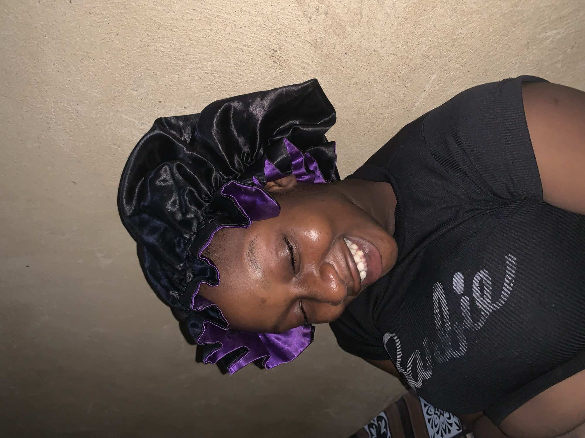 A young Black woman with her eyes closed and mouth open in a wide smile is pictured from the chest up, leaning slightly to her left. She is wearing a black satin bonnet with purple trim and a black ribbed t-shirt. The background is a plain, textured wall. The lighting is dim and appears to be coming from the front and right. There is a hint of a patterned fabric to the left of the woman, partially obscured. The scene is likely indoors.