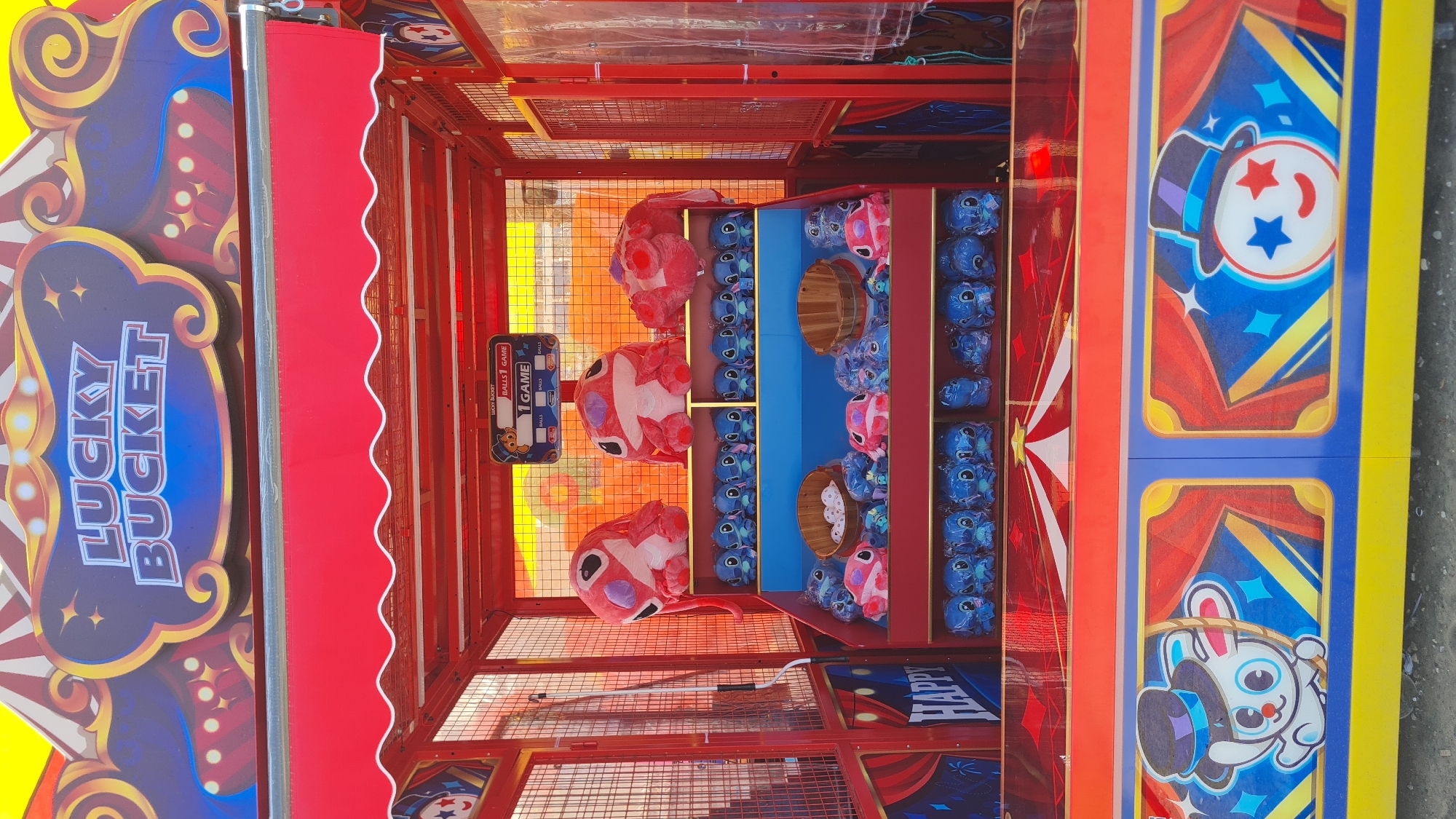 A carnival game booth is visible, featuring a red structure filled with plush toys. Predominantly, there are blue and pink variations of a Stitch-like character. Wooden buckets are also present within the display. On the left side of the booth, a sign reads "LUCKY BUCKET" in large letters. The booth's design incorporates vibrant colors and patterns associated with carnivals. The location is unspecified.