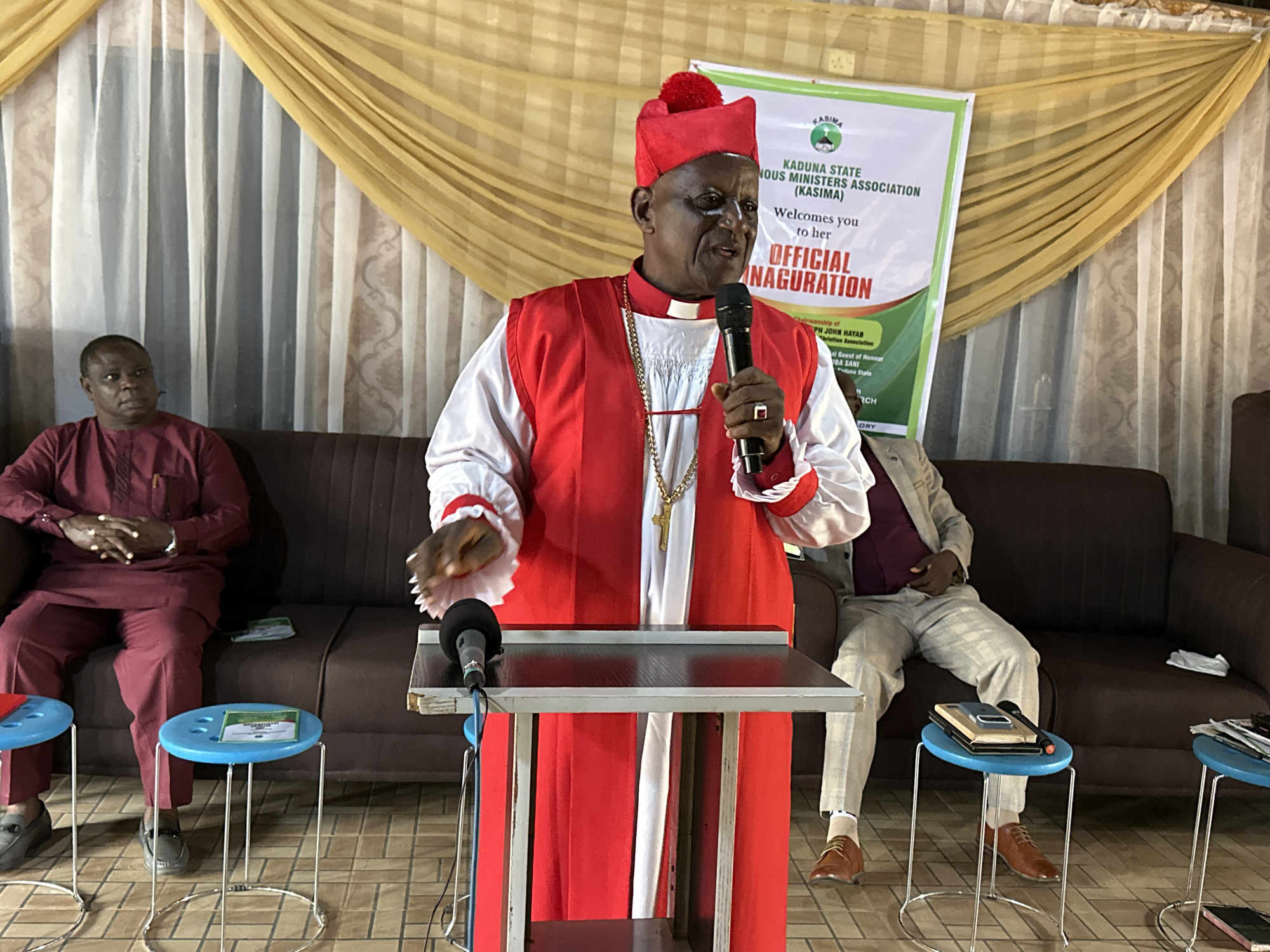 The photograph depicts an indoor ceremony in Barden Yana, Nigeria. In the center, a man in traditional religious attire, including a red skullcap and a red and white robe with a golden cross pendant, speaks into a microphone. He is positioned behind a podium. To his left, a man in a maroon outfit is seated on a brown sofa, with his hands clasped. To the right, another man in a light-colored suit and pants is also seated on the sofa. The background features a draped golden curtain and white curtains with a patterned design. Blue stools are placed in the foreground. A banner with green and white lettering is visible behind the speaker. The lighting suggests an indoor setting with artificial light sources.