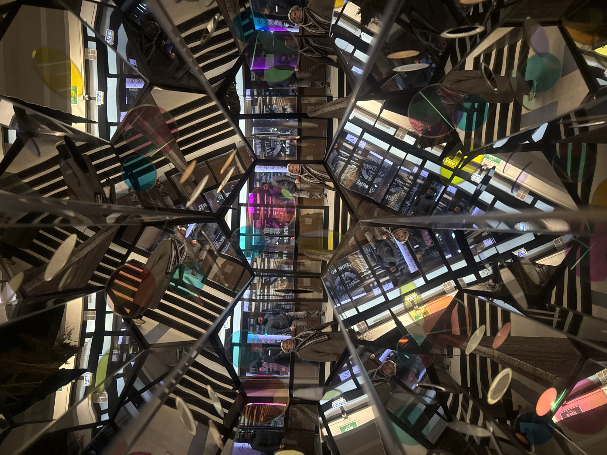 The image presents a complex, kaleidoscopic view from within a mirrored art installation or structure, likely located in Yekaterinburg, Russia. Multiple reflections create a fragmented and repeating pattern of the surrounding urban environment and individuals.
Several people are visible across the mirrored surfaces, dressed in winter attire including puffer jackets, scarves, and beanies, suggesting cold weather. They appear to be moving through or observing the installation. One individual wearing a light-colored beanie and a striped scarf is prominently reflected in various panels.
The scene is illuminated by artificial lights, indicating it is likely evening or night. The interior of the installation features striking black and white striped patterns on some surfaces. Numerous translucent, colored disks, in hues of yellow, blue, green, pink, and orange, are suspended or projected, contributing to the abstract, kaleidoscopic effect.
Visible text, both in Cyrillic and English, includes "ВЫХОД" and "EXIT" on green signs, along with other fragments such as "GROTT", "НАСТОЯЩЕЕ" (Real), "САМЫЕ" (The Most), "ДОБРО" (Good), and "ПЛОВ" (Plov), which is a dish often associated with restaurants. A numerical sequence "10098" is also visible. These texts, especially the Cyrillic script, are consistent with the given location context of Yekaterinburg, Russia, suggesting reflections of nearby commercial establishments. The overall impression is an immersive and visually stimulating experience.