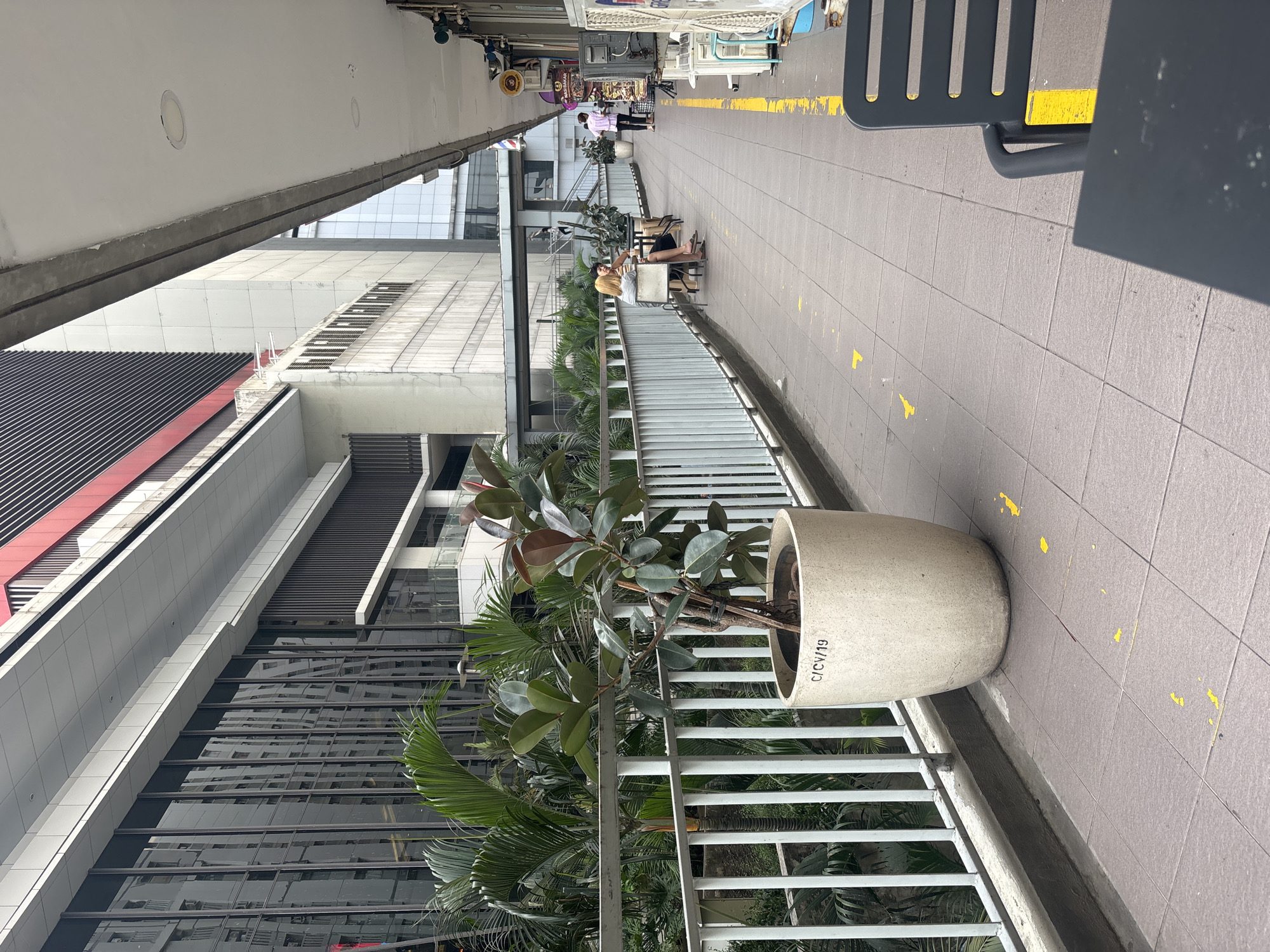 An outdoor walkway, likely in a commercial or pedestrian area of North Jakarta, Indonesia, features a tiled floor with yellow directional markings. To the left, a white railing runs along the walkway, separating it from a lush, green garden with palm trees and other tropical foliage. A large, light-colored planter contains a potted plant with broad, dark green leaves. In the mid-ground, two individuals are seated at a small table with chairs, appearing to be engaged in conversation. Further down the walkway, a person in a white shirt and dark pants walks away from the camera, and several shops or stalls are visible on the right, with one sign reading "Nasi Bakar." Air conditioning units are mounted on the exterior walls of the buildings on the right. The background is dominated by modern, multi-story buildings with reflective glass facades.