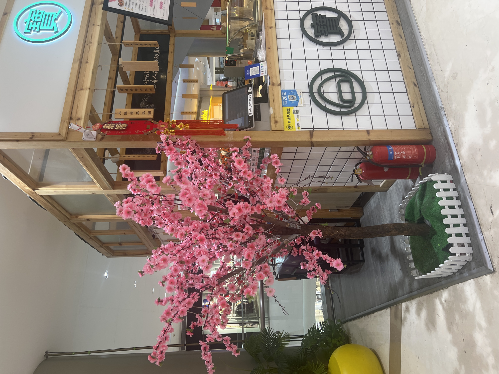 The image depicts an indoor setting, likely a commercial establishment in Liandu District, China. A large, artificial pink cherry blossom tree with a brown trunk stands prominently in the foreground, its branches extending across the frame. The tree is surrounded by a small white picket fence with artificial green turf. To the right of the tree is a food stall or counter made of light-colored wood and white tiled walls. Above the counter, a glowing blue sign with a Chinese character is visible. The counter area features a POS system with a touch screen monitor, a QR code for payment, and various food-related signage in Chinese. Two red fire extinguishers are positioned to the left of the counter, near the base of the cherry blossom tree. In the background, a shop interior with displays and lighting can be seen through a glass partition, suggesting this scene is located within a mall or shopping center. The overall environment appears clean and well-lit. No people are clearly visible in the immediate vicinity of the stall.