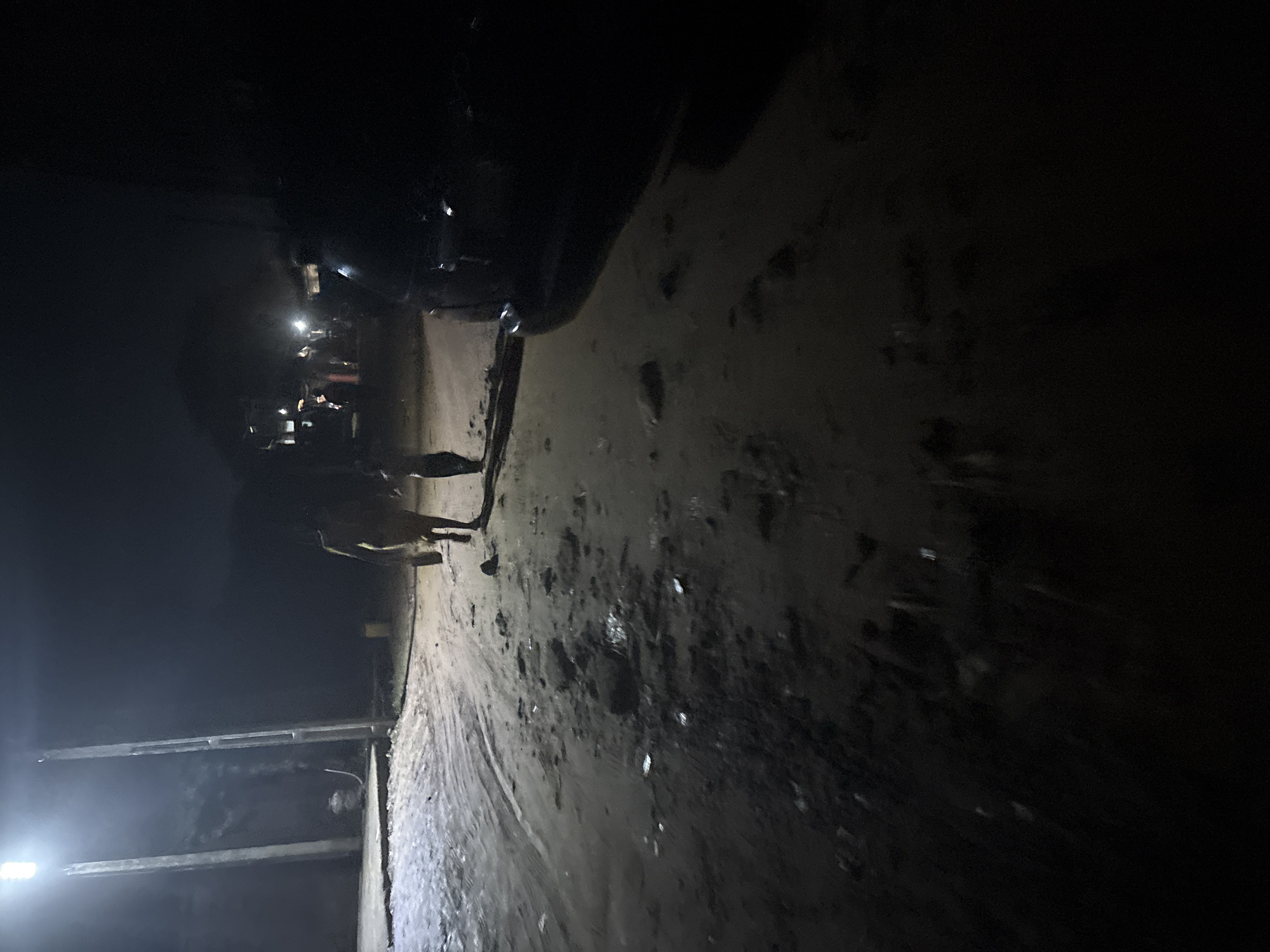 The image depicts a dimly lit outdoor scene, likely at night, on an unpaved, wet surface in Onitsha, Nigeria. A tall, illuminated pole with a bright light stands on the left side of the frame, casting a strong glow on the ground and a concrete path. In the middle ground, a person, appearing to be an adult, is walking away from the camera, holding a light-colored object in their left hand. A smaller figure, possibly a child, walks closely behind them. Further in the background, other figures and what appear to be structures with lights are faintly visible. To the right of the frame, the rear of a dark-colored vehicle is partially visible. The ground is uneven, with puddles of water and scattered debris. The overall impression is one of a rural or semi-urban environment at night, illuminated by artificial light.