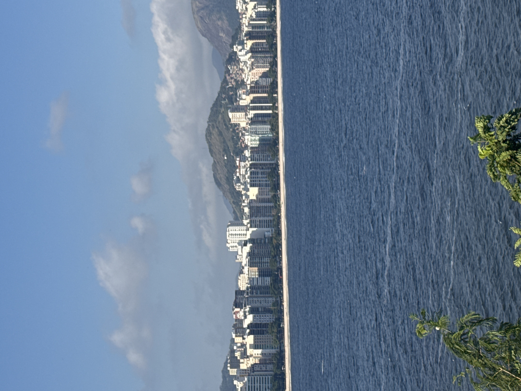The image depicts a panoramic view of the coastline of Niterói, Brazil, facing the Guanabara Bay. In the foreground, the deep blue waters of the bay stretch across the frame, with subtle ripples indicating a gentle breeze. To the left and right, the lower portions of lush green foliage are visible, framing the scene.
Dominating the midground is the city's distinctive skyline, characterized by a long stretch of beachfront lined with numerous high-rise apartment buildings. These structures vary in height and architectural style, with many featuring balconies and distinct window patterns. The buildings are primarily painted in shades of white and pastel colors, creating a bright and modern urban landscape. Behind the city, rolling green hills and the rounded peaks of mountains form a dramatic backdrop. The sky above is a clear, light blue, with scattered, wispy white clouds that suggest fair weather. The overall impression is one of a vibrant coastal city bathed in bright daylight.