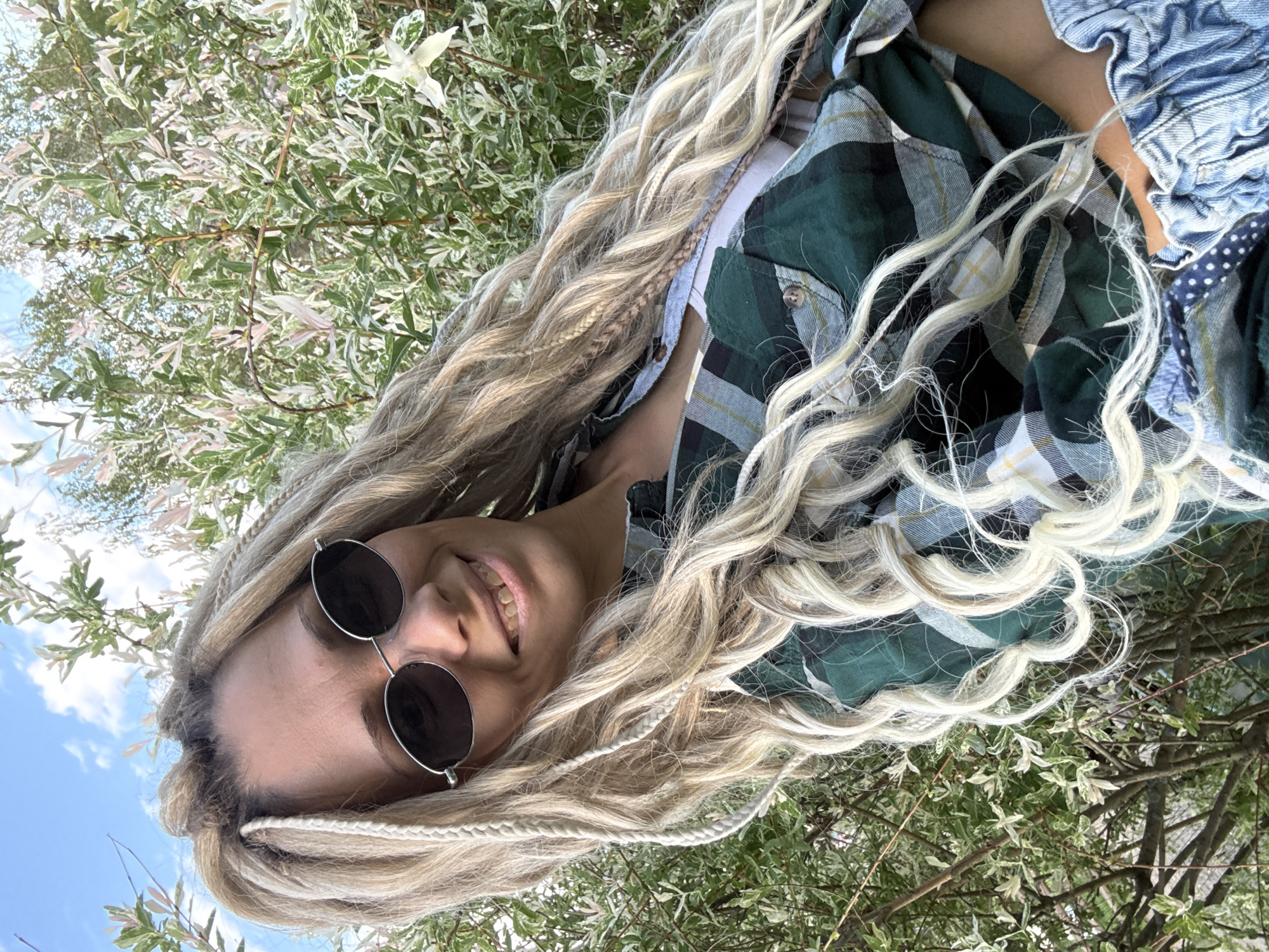 A young woman with blonde hair, wearing round sunglasses and a green and white plaid shirt over a white top, is smiling at the camera. She is outdoors, with a tree with white and green leaves in the background and a blue sky visible. The location appears to be a park or green space. The setting is not directly identifiable as Rusanovo, Russia based on the visual information.